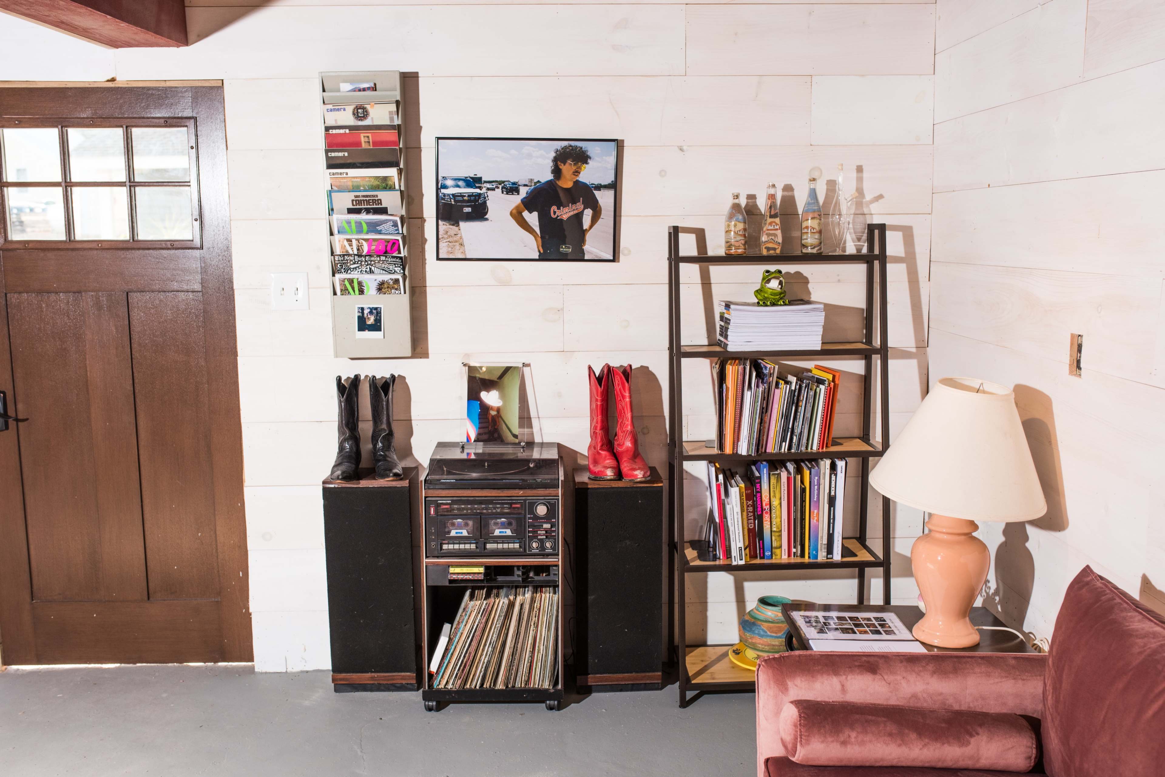 A room corner with a wooden bookshelf, a record player, a pair of black and red boots, a lamp, and a framed photo on the wall.