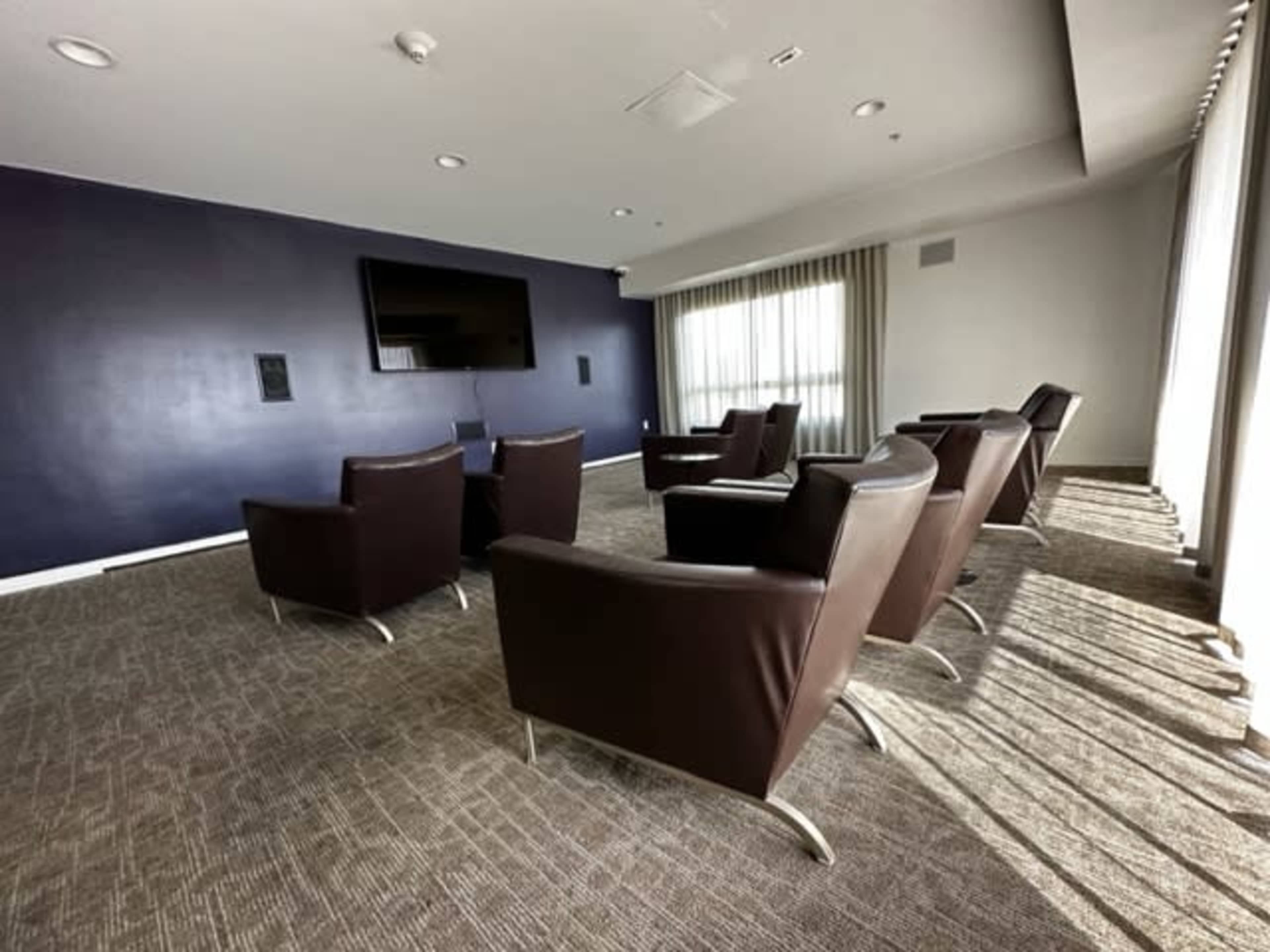 A room with five brown upholstered chairs arranged facing a wall-mounted TV, with large windows covered by sheer curtains letting in natural light.