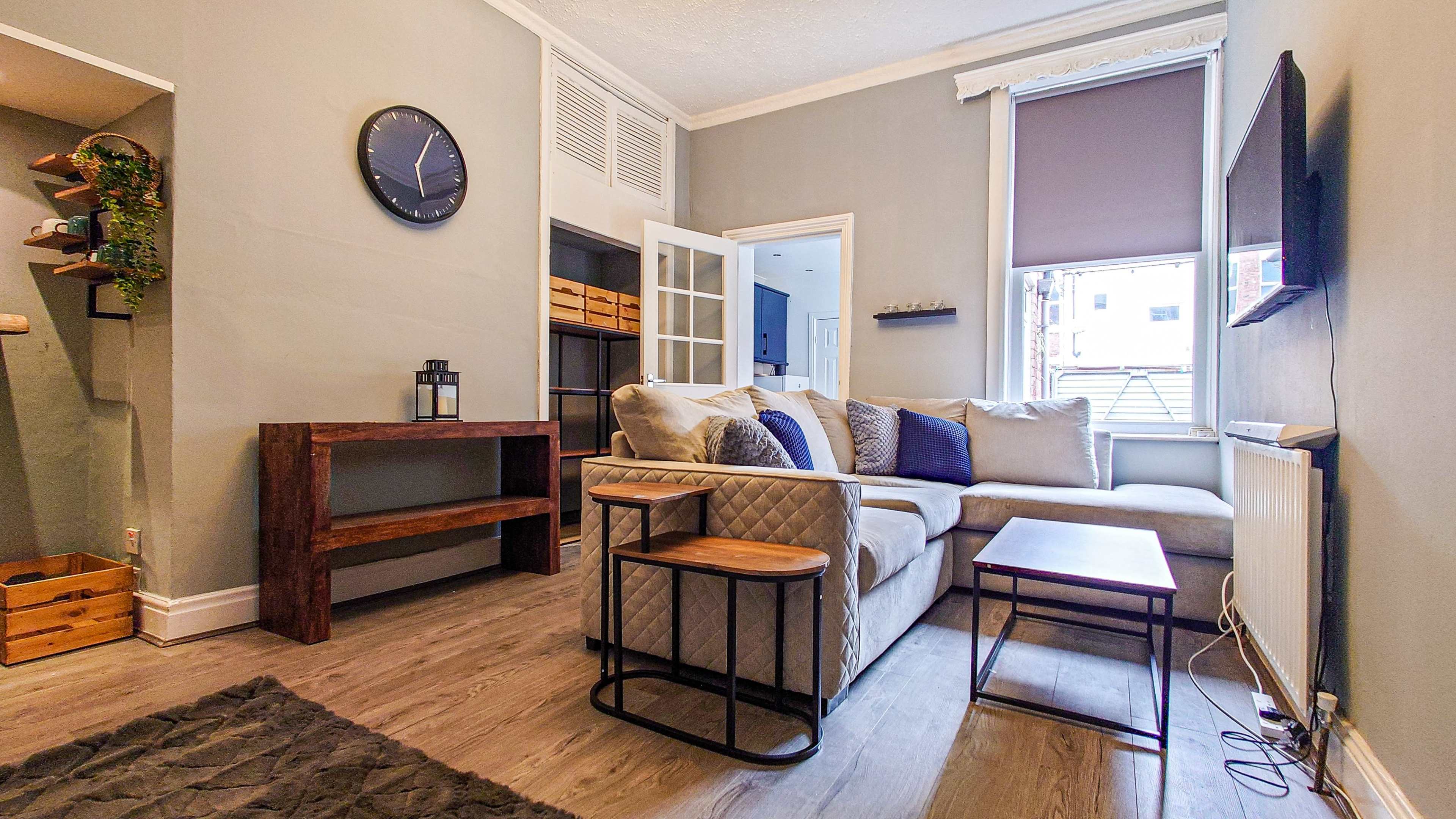 A small living room with a beige sectional sofa, a wooden coffee table, and a wall-mounted television beside a window.