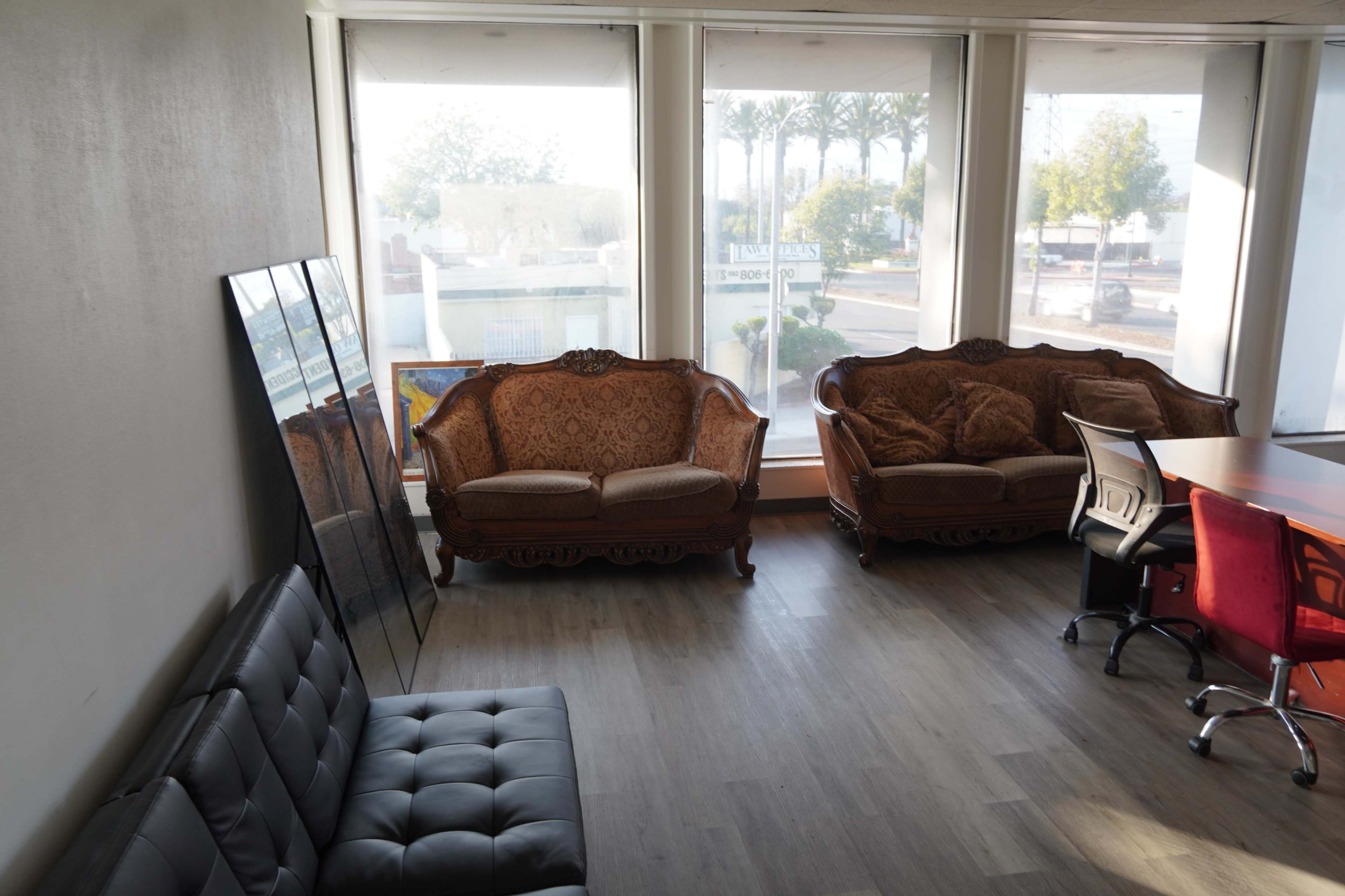 The image shows a spacious office area featuring two ornate brown couches and a black tufted sofa, with large windows allowing natural light to enter.