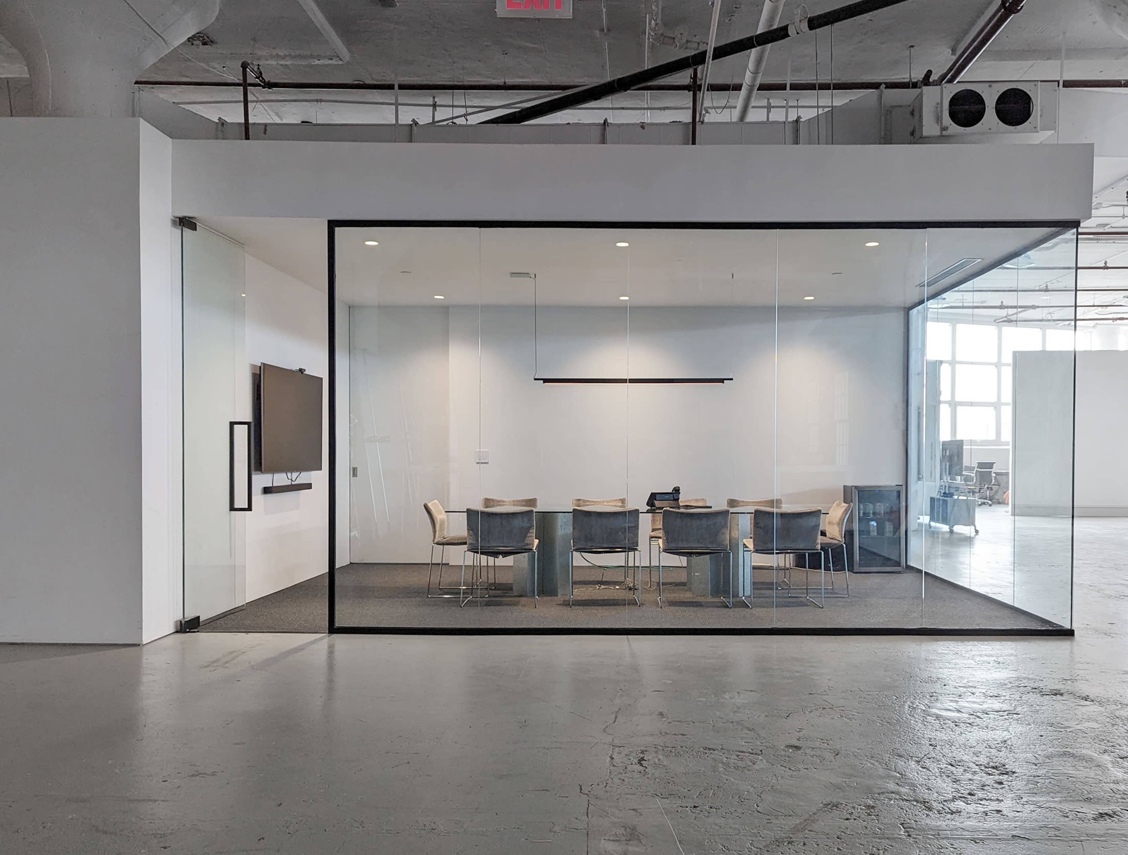 The image shows a modern conference room enclosed in glass, featuring a long table surrounded by chairs and a wall-mounted screen.