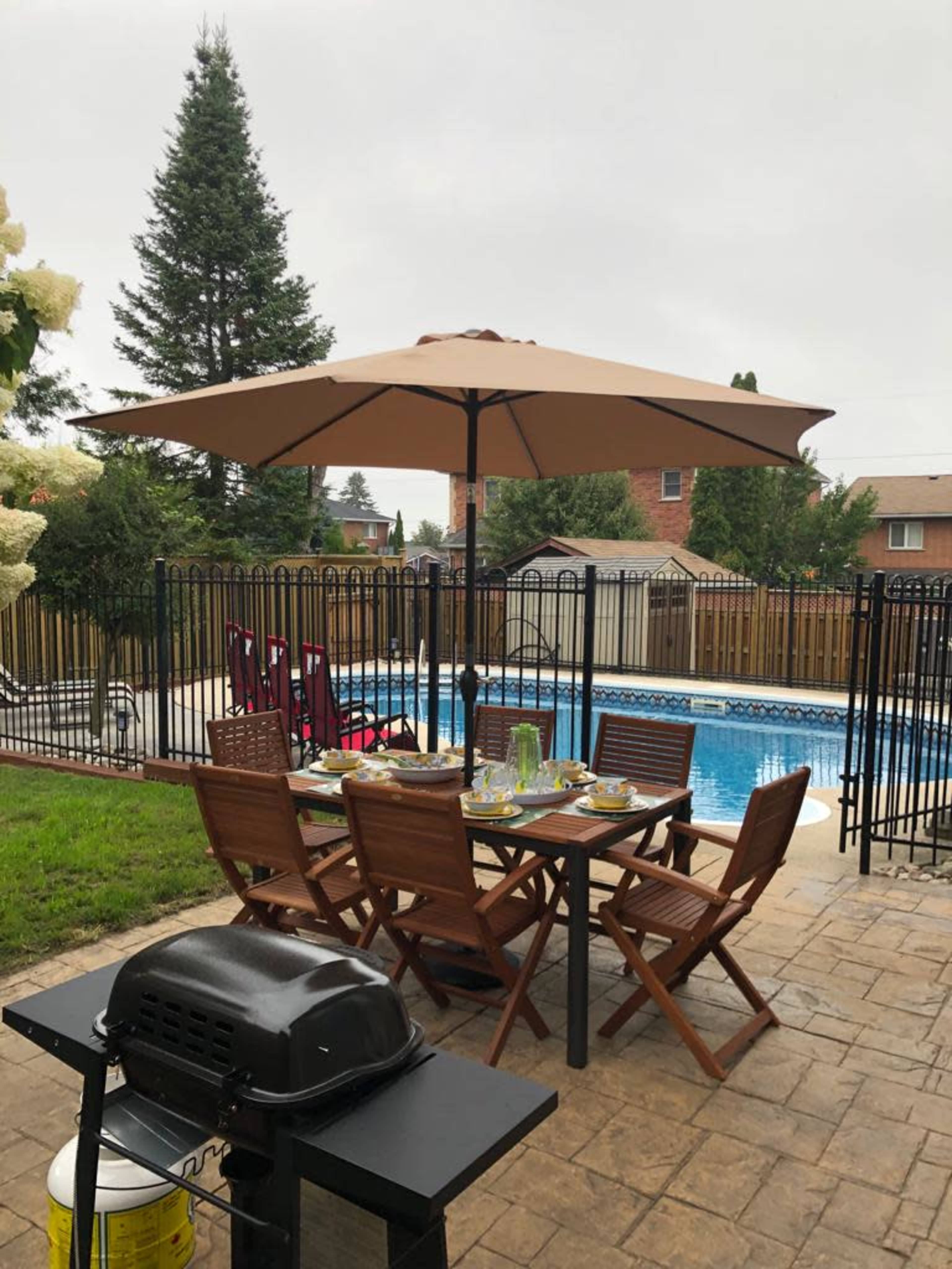 A backyard patio features a table set for dining under an umbrella, overlooking a fenced swimming pool.