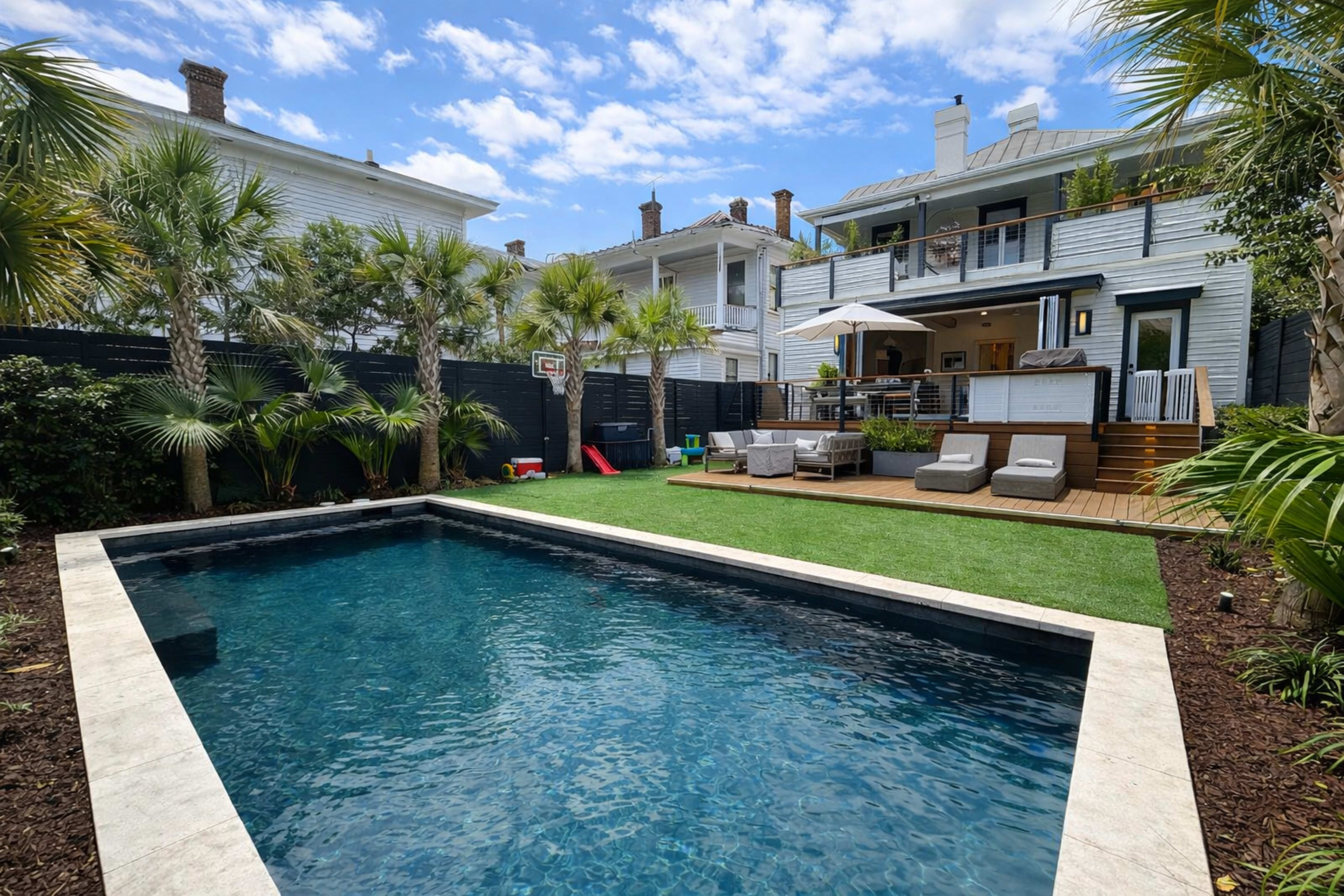The image shows a backyard with a swimming pool, surrounded by palm trees and featuring a two-story house with a deck, patio furniture, and a basketball hoop.