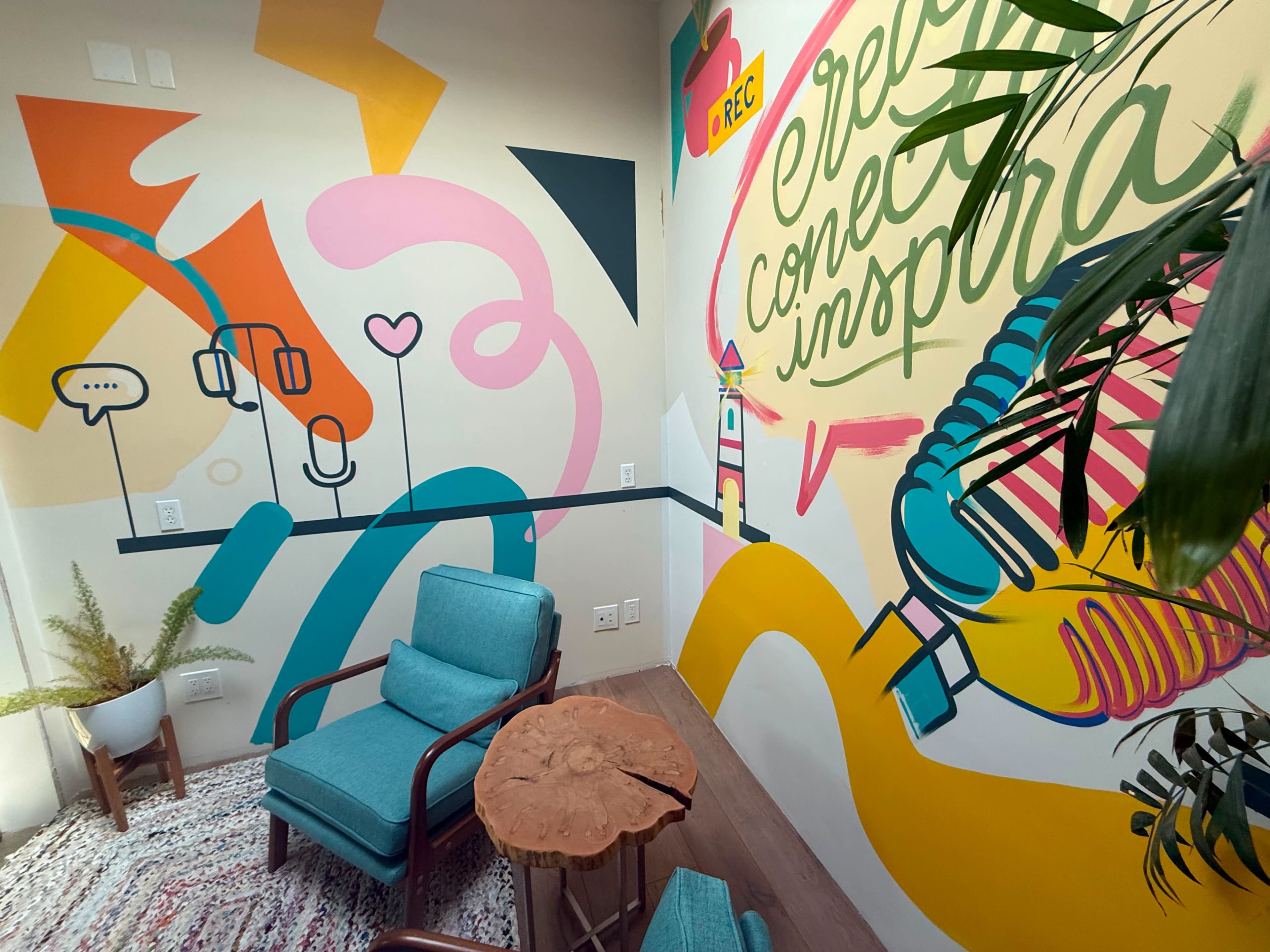The image shows a colorful room with vibrant wall murals, two blue chairs, and a wooden table.
