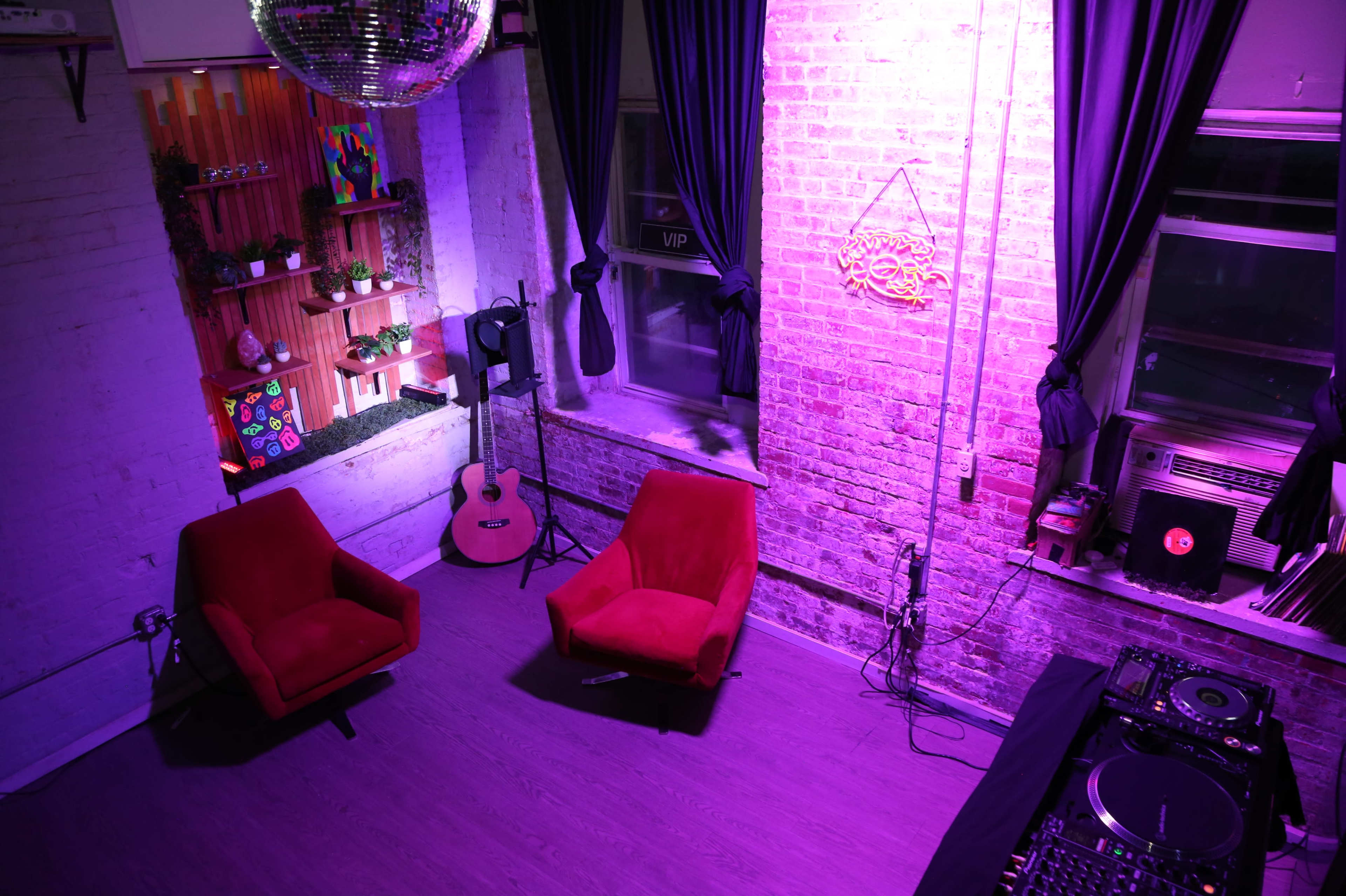 The image shows a brightly lit room with two red chairs, a guitar, a DJ setup, and plants on a shelf against a brick wall.
