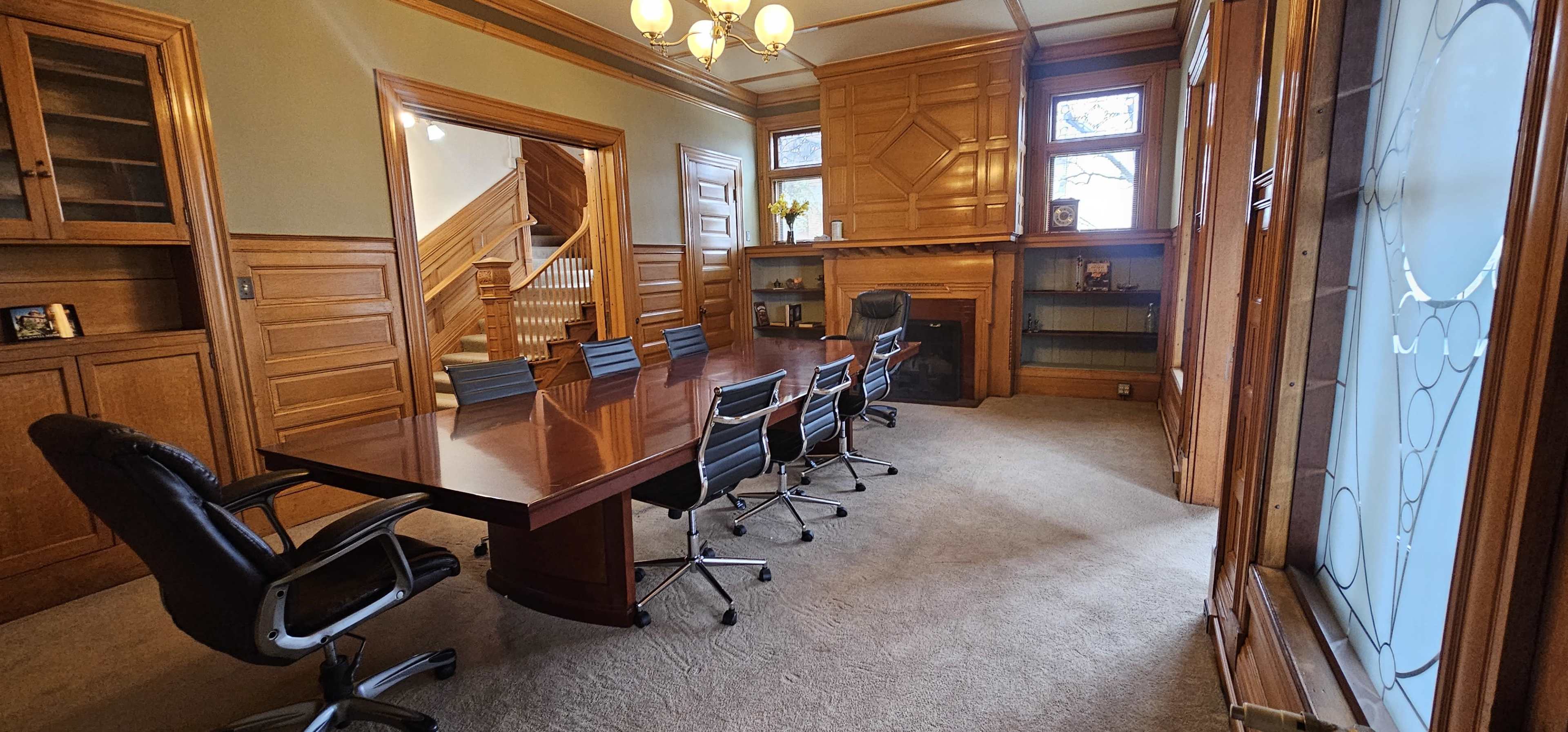 A conference room features a long wooden table surrounded by black chairs, with wooden paneling and a fireplace along one wall.