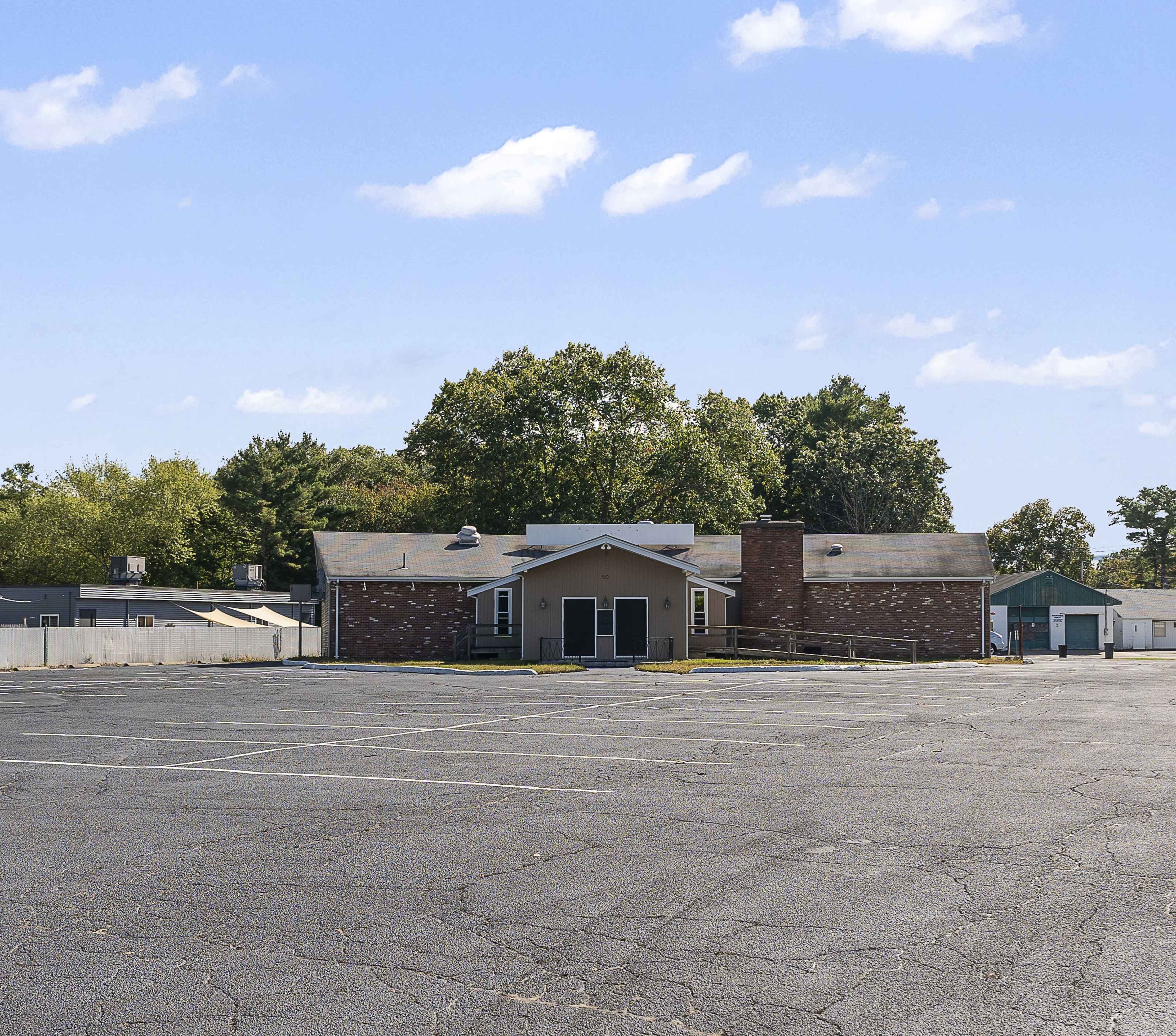 A large, empty parking lot stretches in front of a single-story brick building with a central entrance and a chimney, surrounded by trees and additional structures in the background.