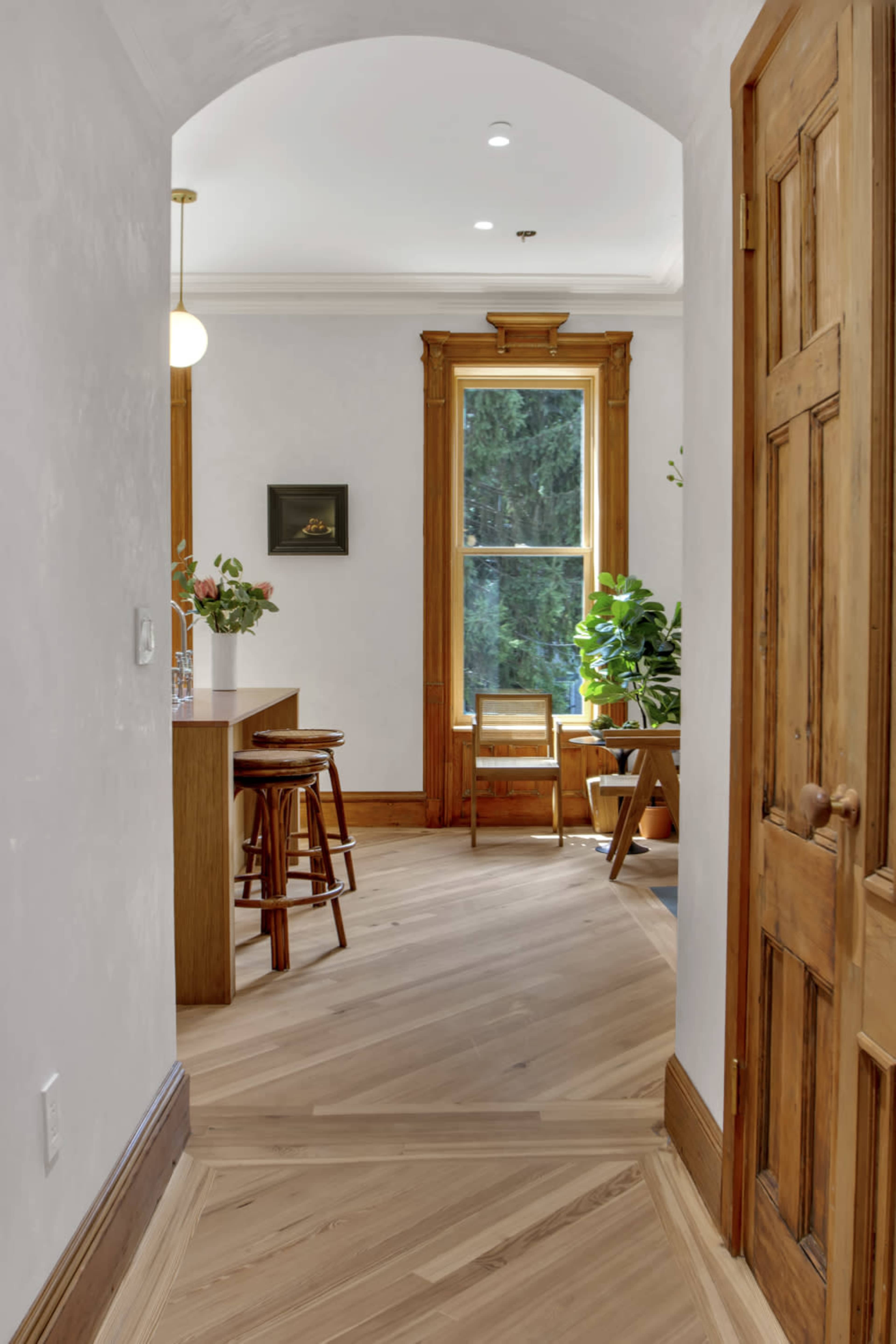 A hallway leads to a sunlit room with wooden furniture and plants, featuring a large window and a bar area.