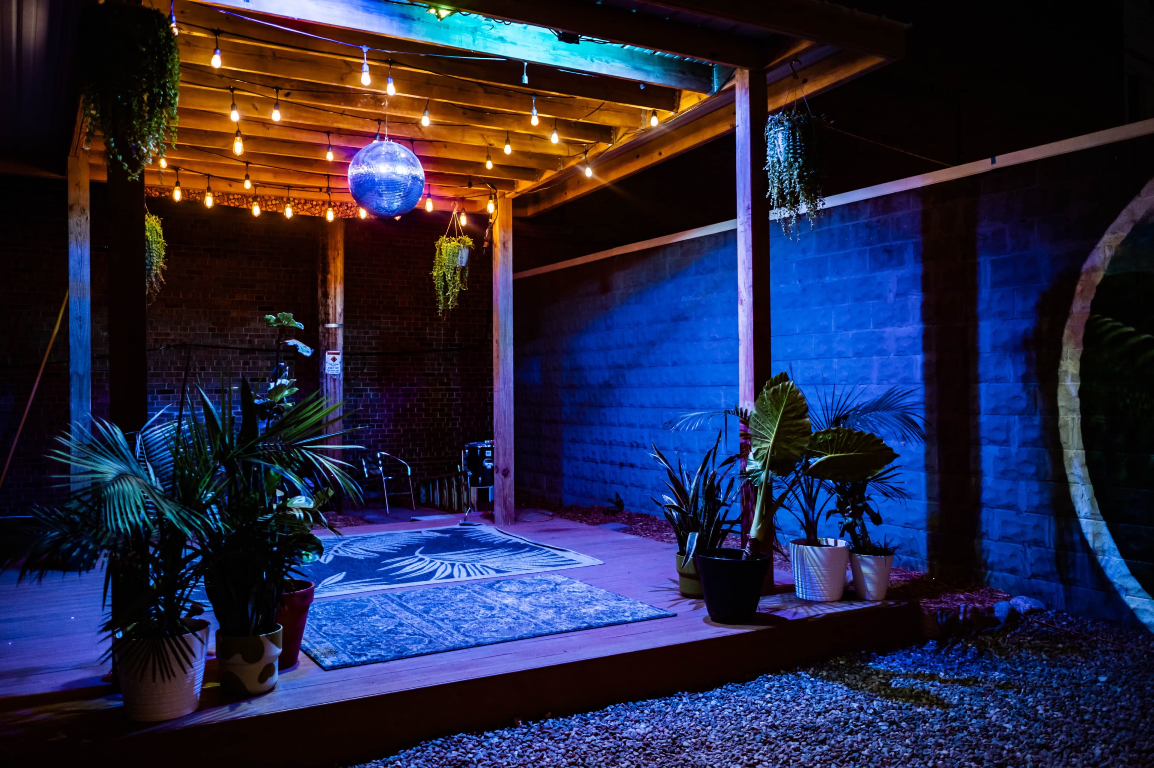 A cozy outdoor area with wooden beams, colorful lights, a disco ball hanging from the ceiling, and potted plants arranged around a patterned rug on a gravel surface.