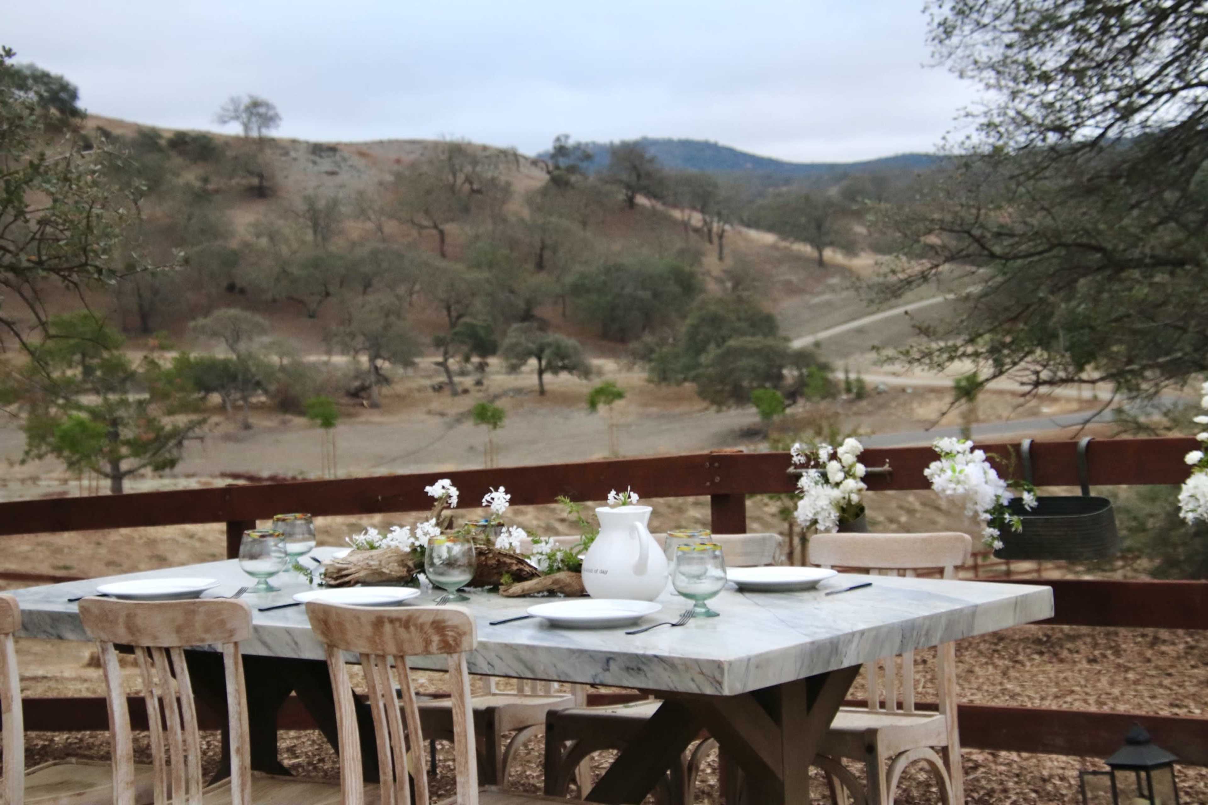 A rustic outdoor dining table is set with plates and glasses, surrounded by an arid landscape with sparse trees and hills in the background.