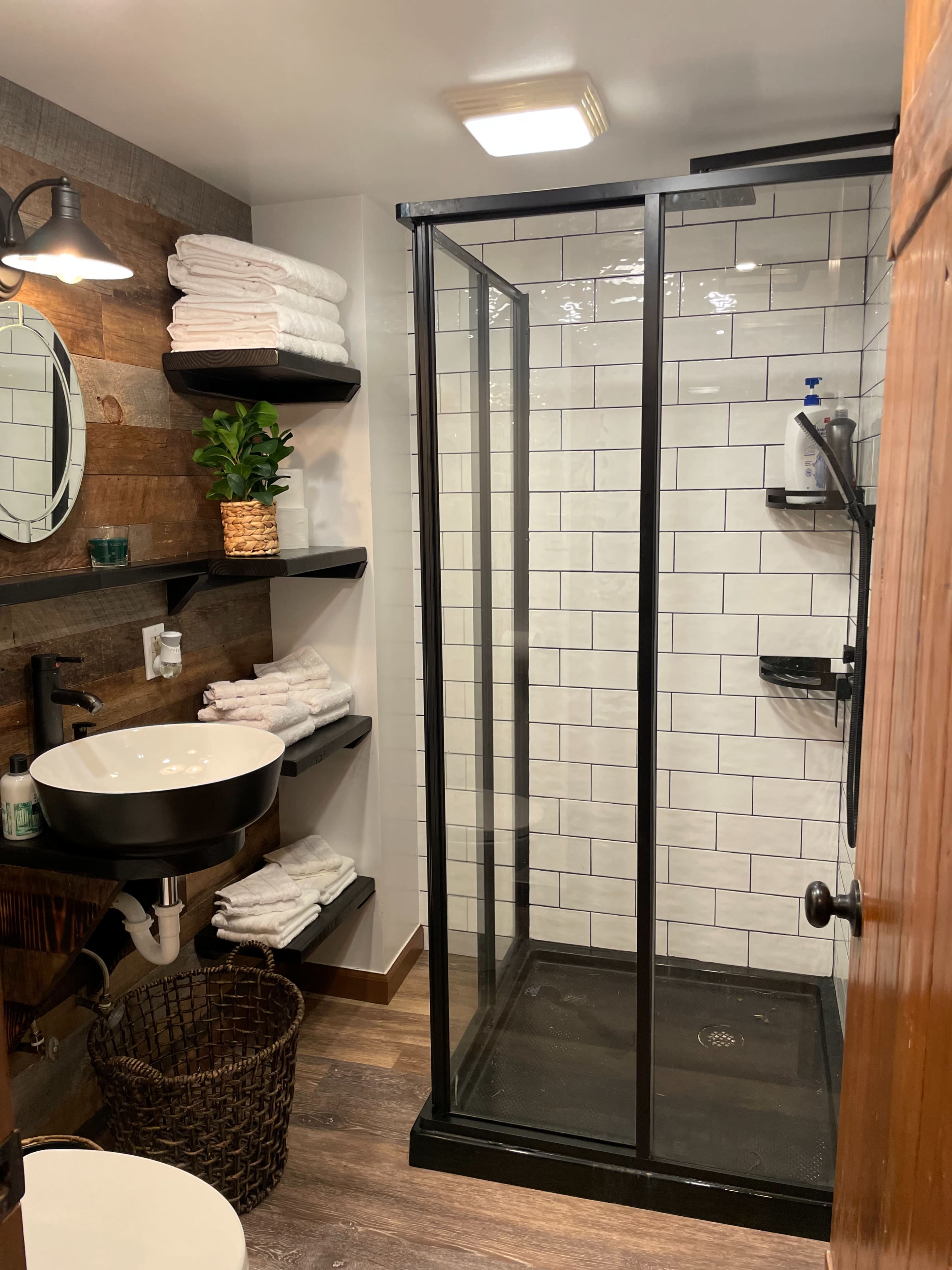 A bathroom featuring a glass-enclosed shower, a round mirror, a sink with a black basin, and neatly arranged towels on open shelves.