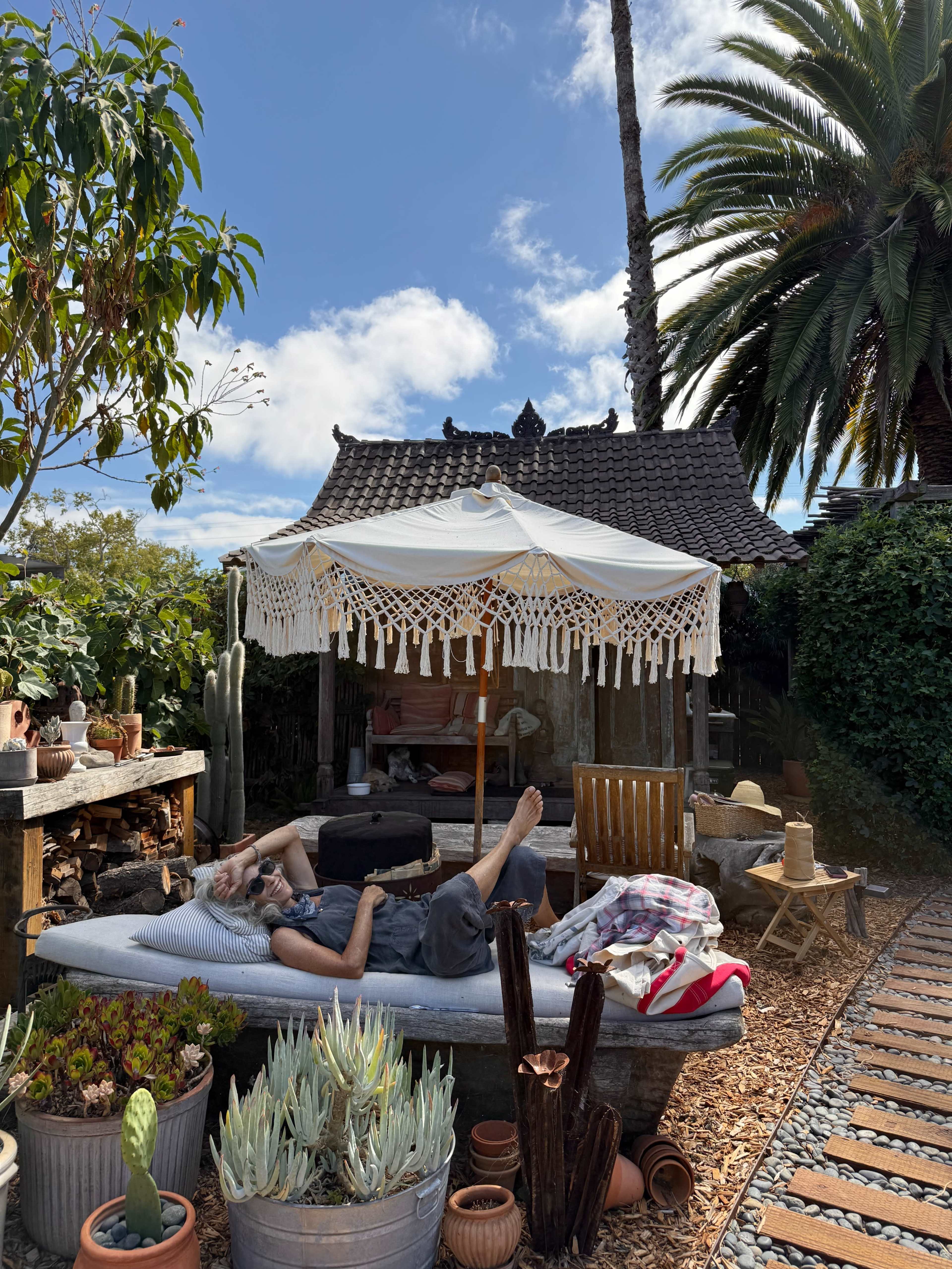 Two people relax on a large cushion under a shaded structure in a garden filled with various plants and pots.