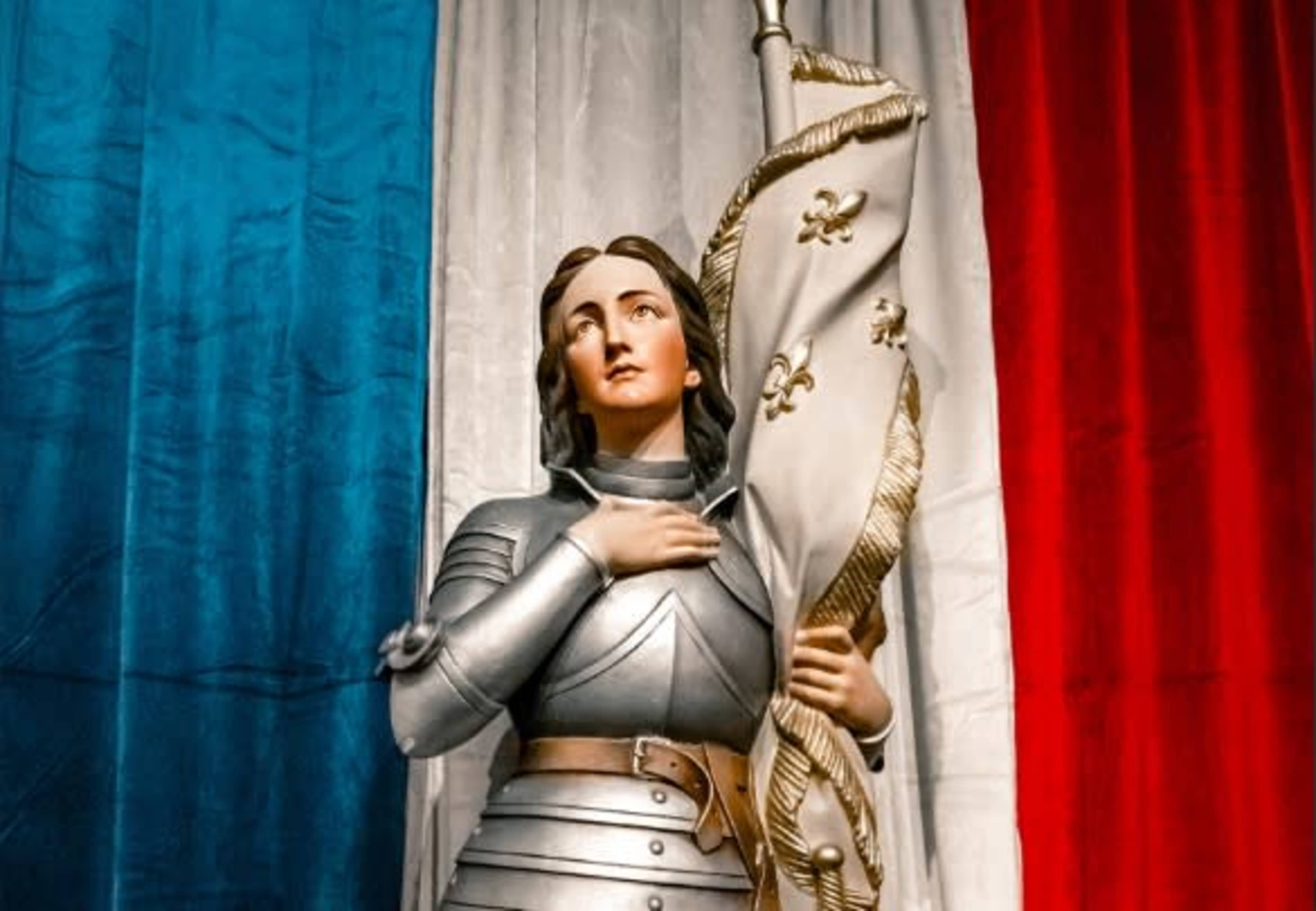The image shows a statue of a armored female figure holding a flag, set against a backdrop of blue, white, and red drapes.
