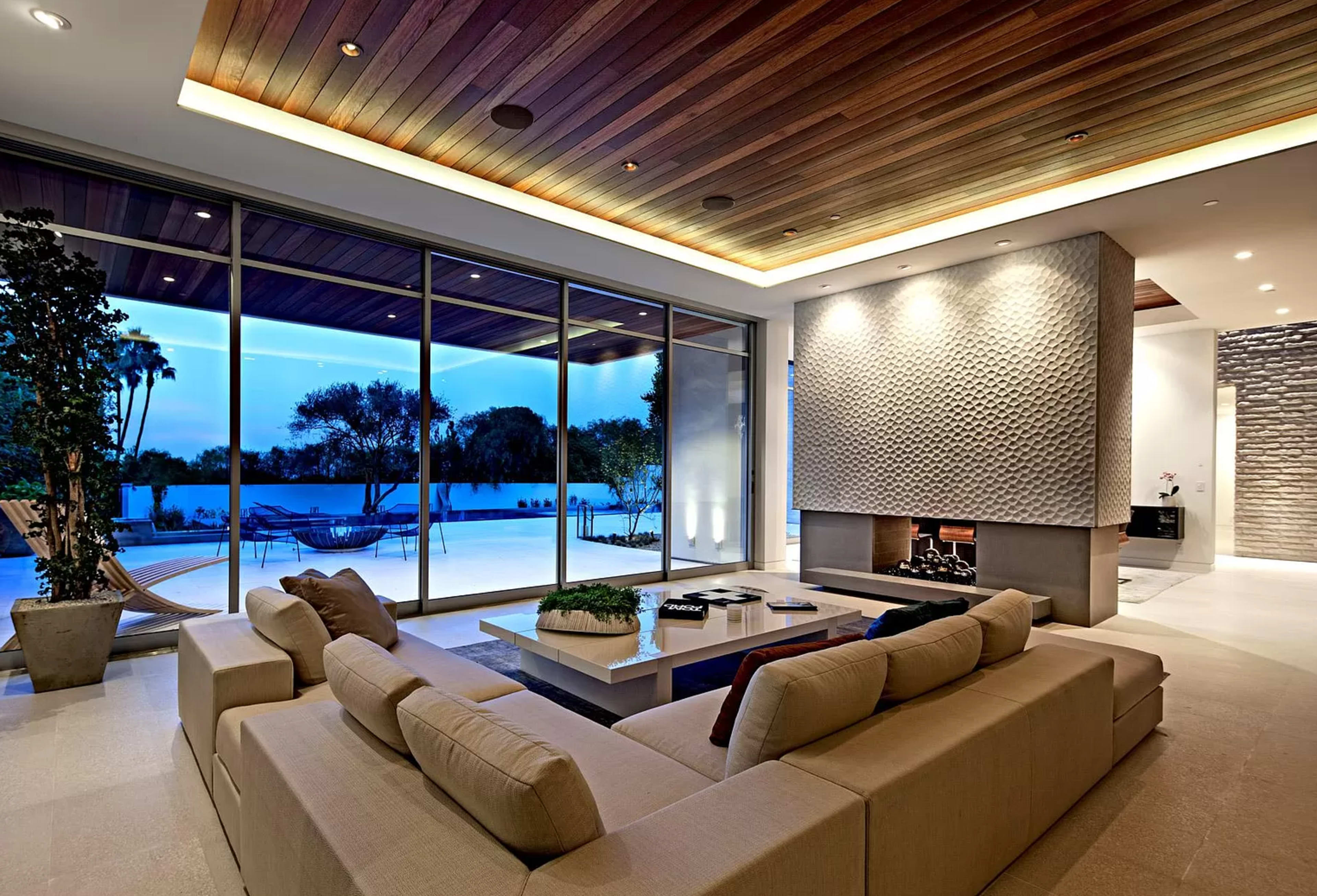 The image features a modern living room with large glass windows overlooking a swimming pool, a light-colored sofa set, and a textured stone accent wall.
