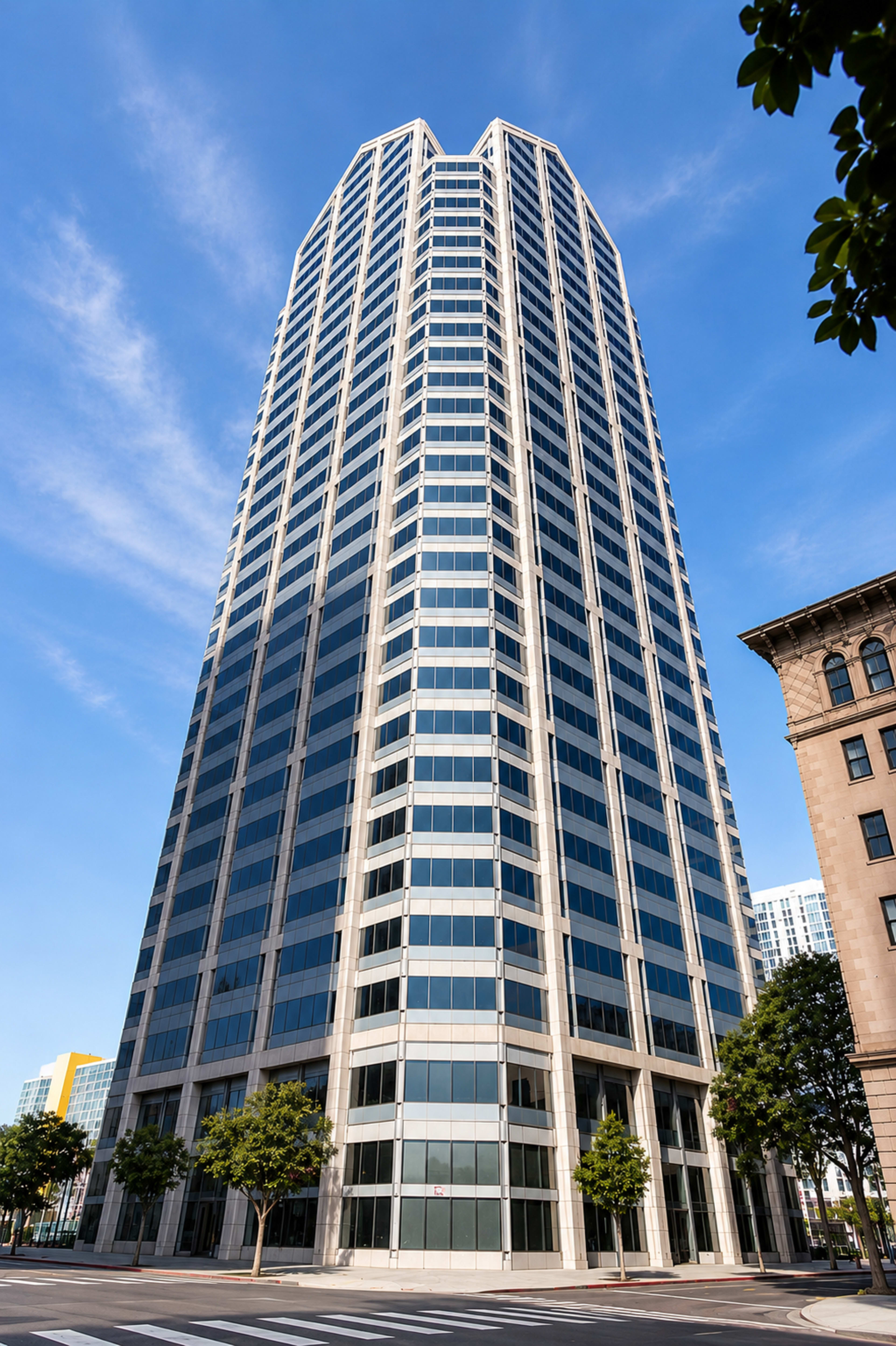 The image shows a tall, modern glass skyscraper with a distinctive angular roof, surrounded by smaller buildings and trees in an urban setting.