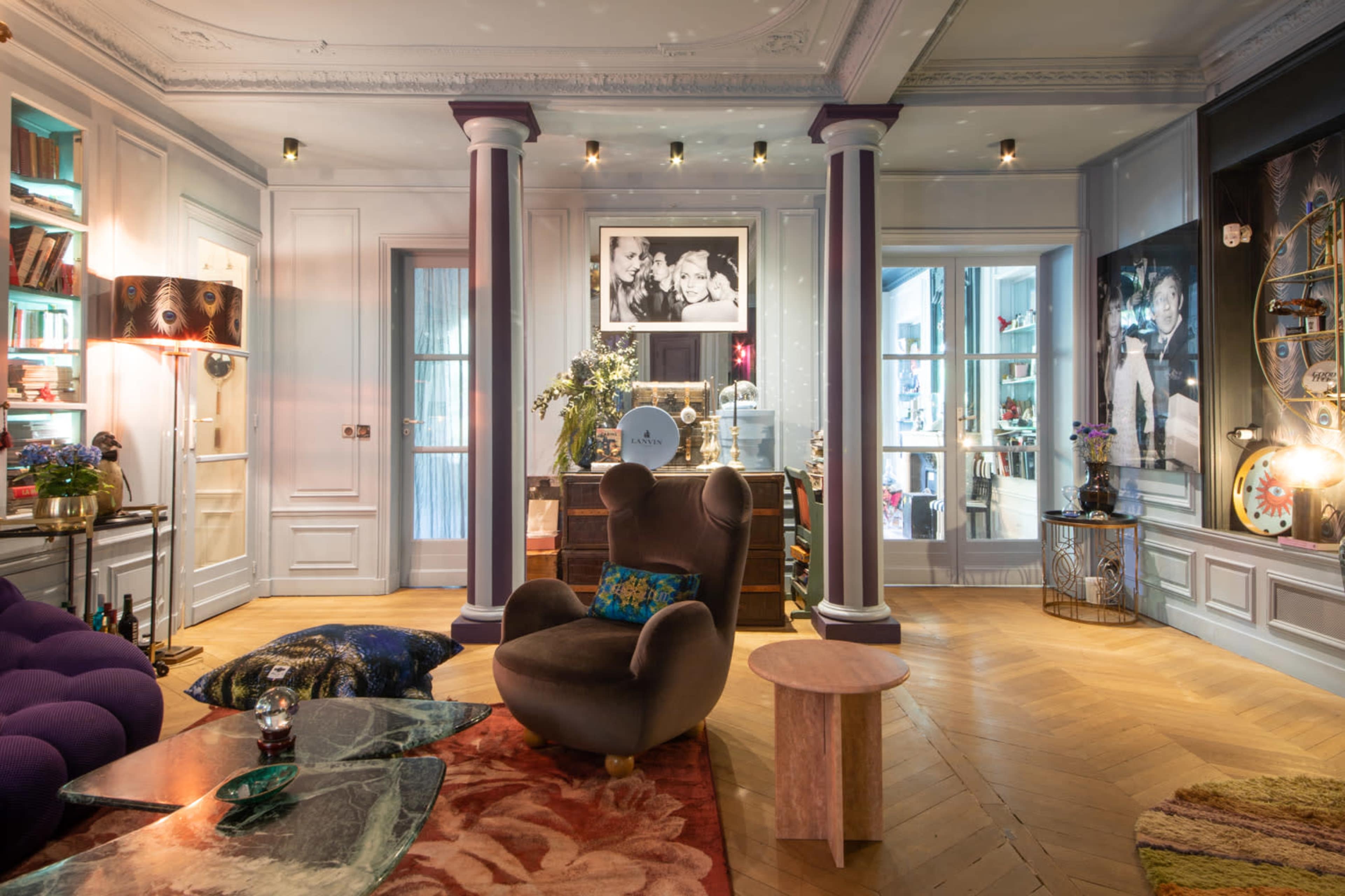 The image shows a stylish living room featuring a purple sofa, a gray armchair, a round coffee table, and various decorative elements, including photographs and plants.