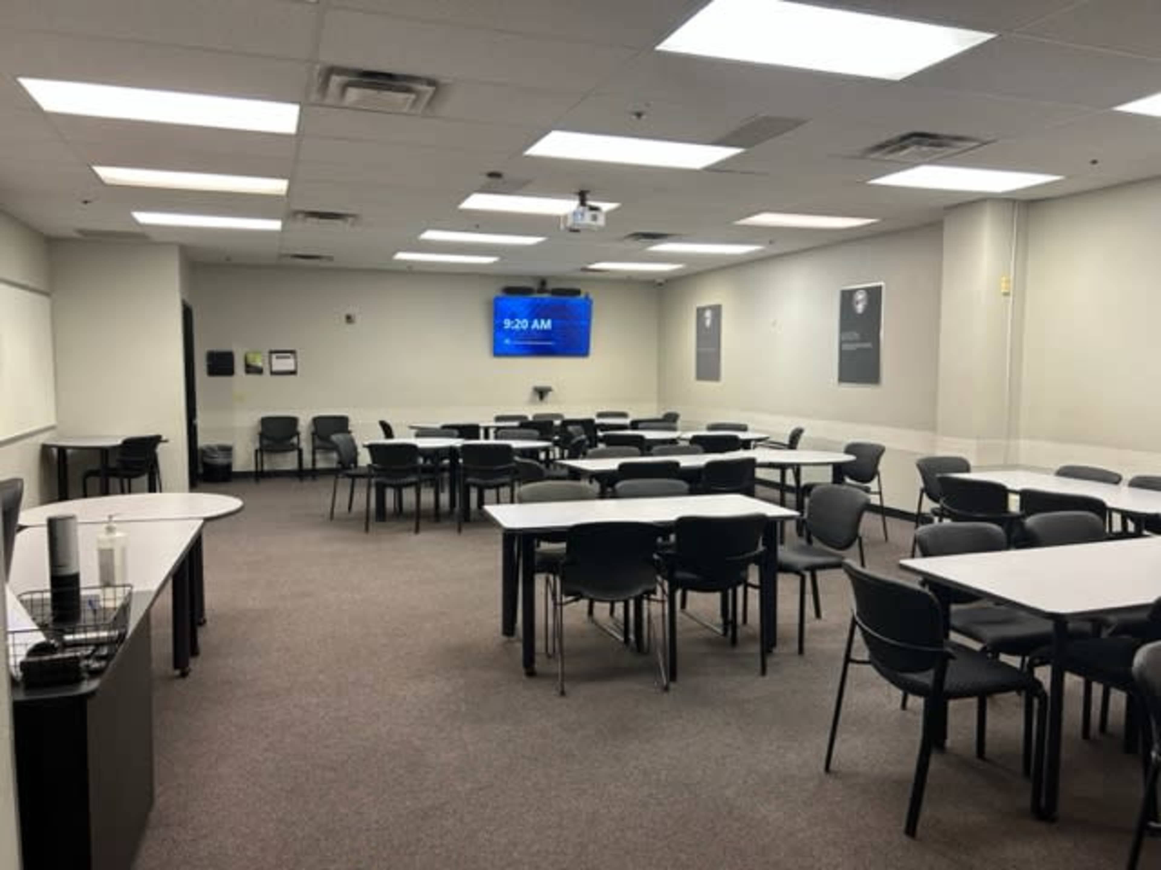 A spacious classroom is set up with multiple rows of tables and chairs, featuring a clock display showing 9:20 AM on a wall-mounted screen.