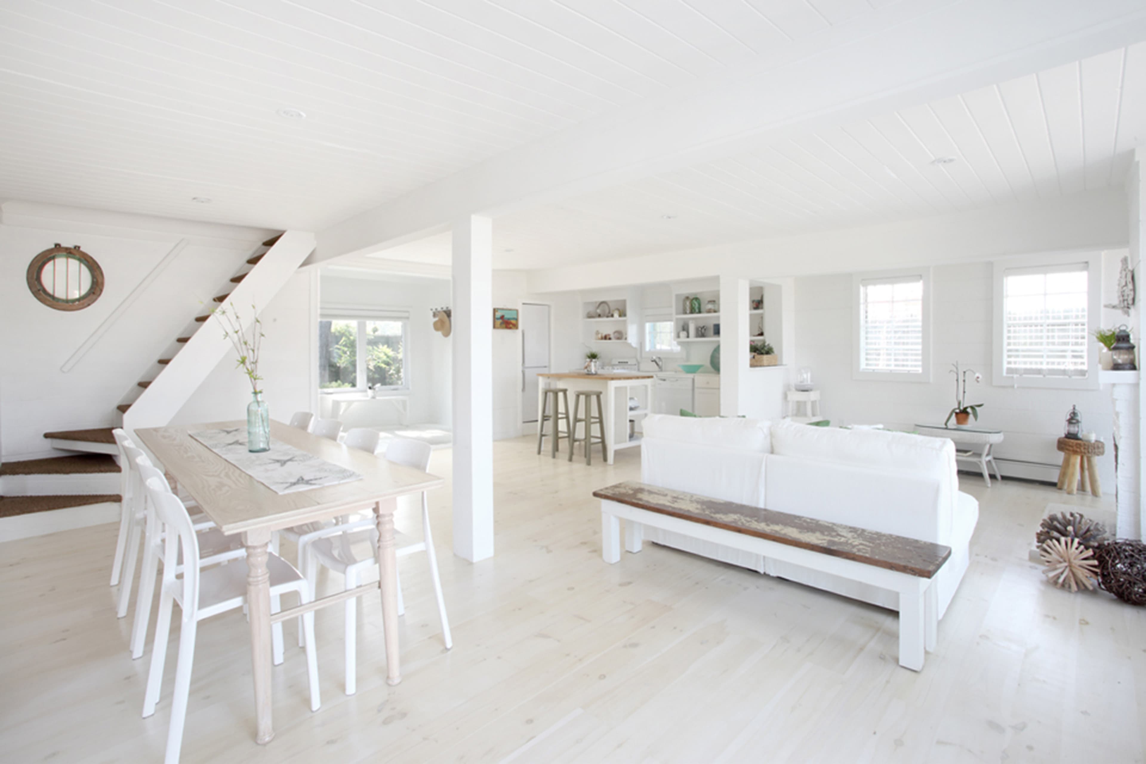 A bright, open living space with a white color scheme, featuring a dining area with a wooden table and chairs, a sitting area with a white sofa, and a staircase in the background.