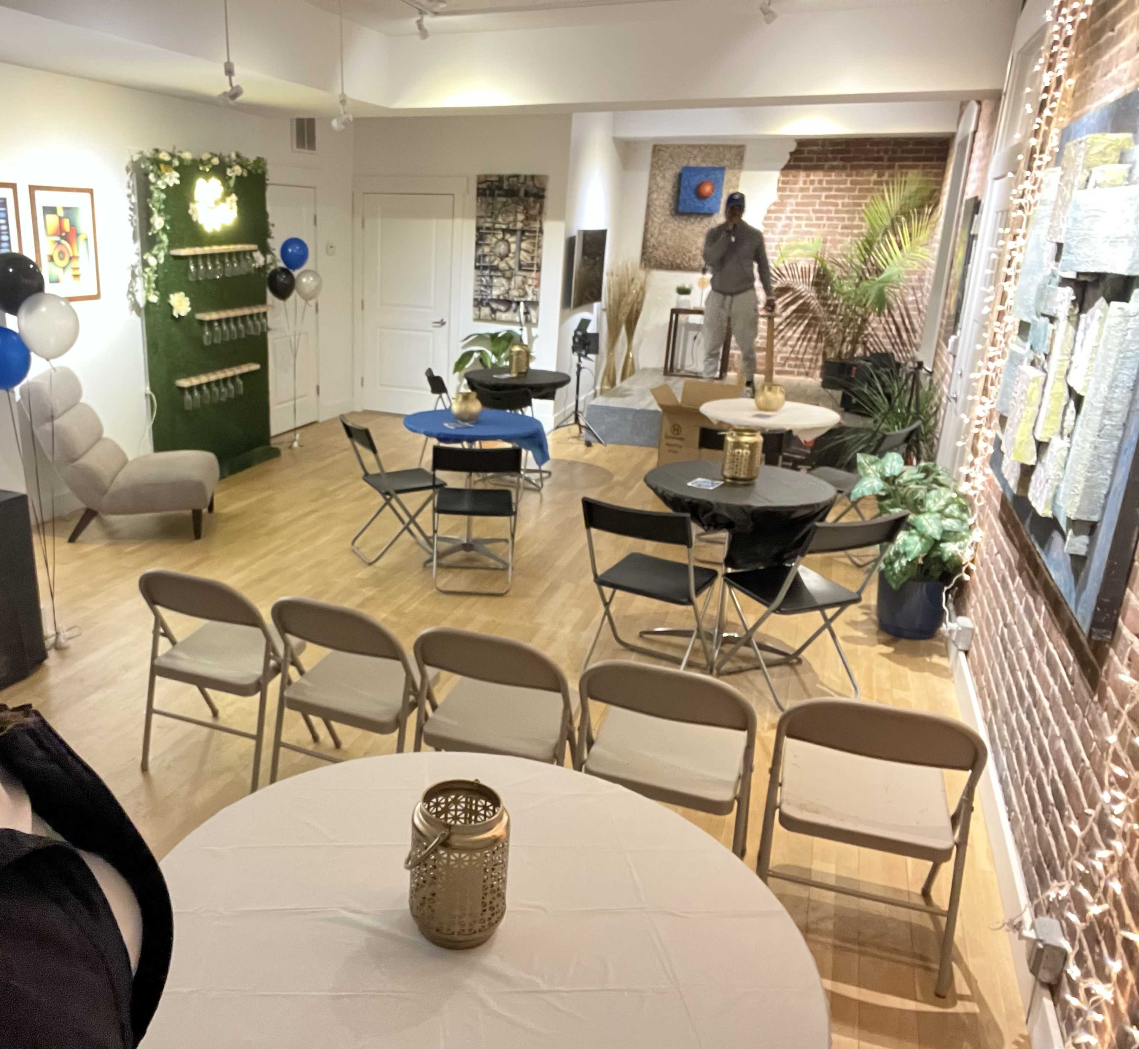 The room features several tables and chairs arranged for an event, with a green wall adorned with decorations, and a person setting up in the background.