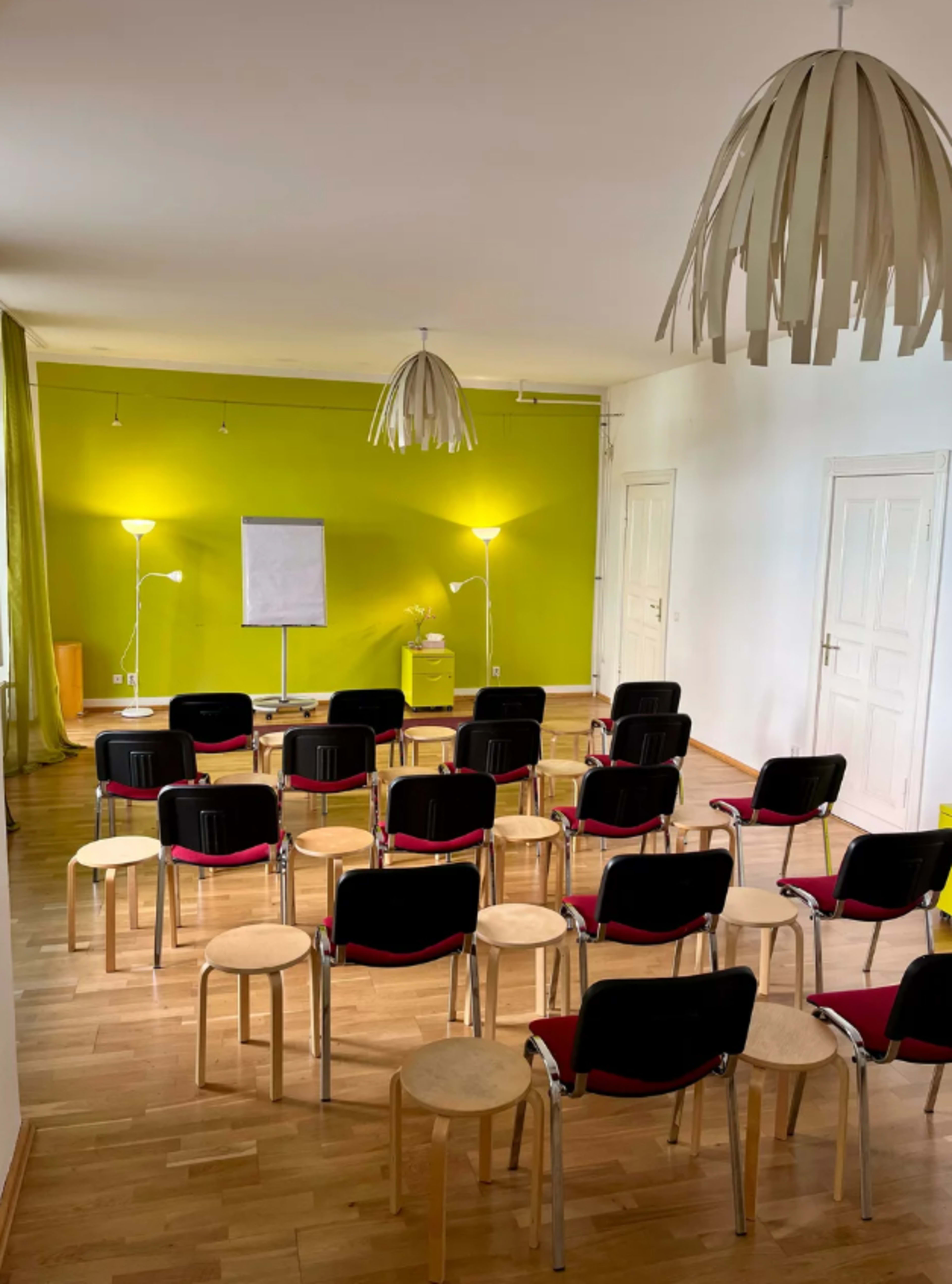 The image shows a spacious room with a green accent wall, featuring rows of empty black and wooden chairs facing a stand-up whiteboard and two floor lamps.