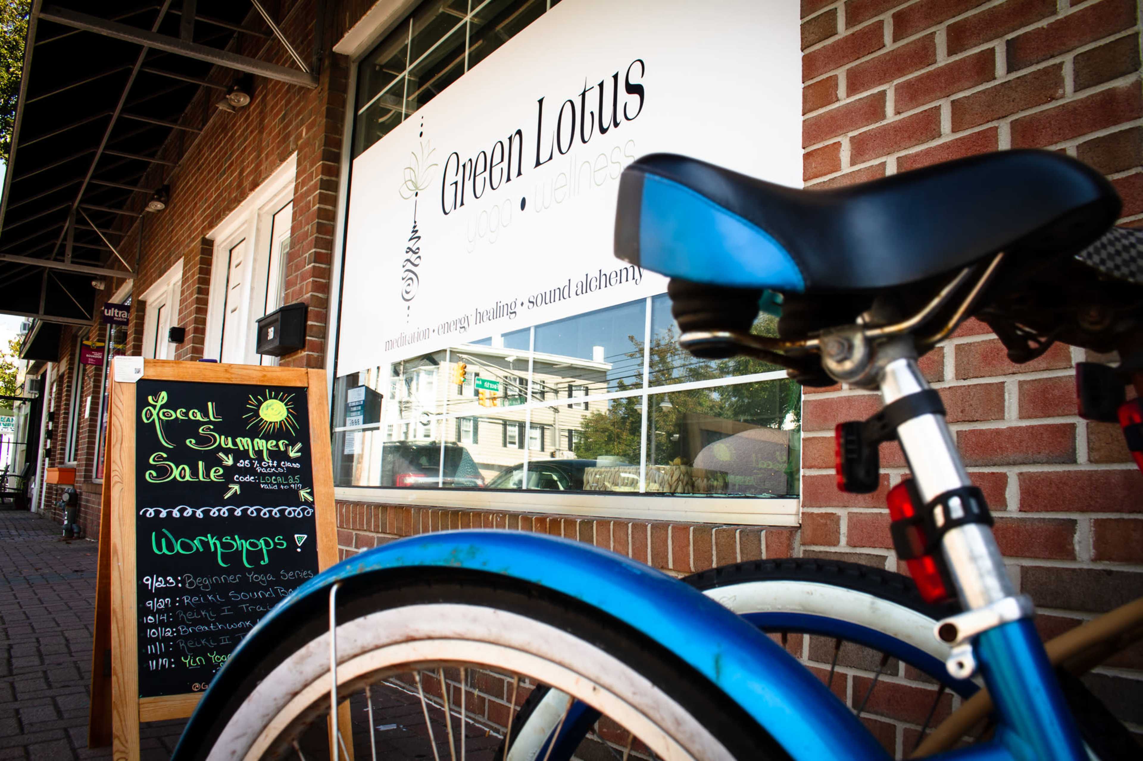 A blue bicycle is parked in front of a storefront with a sign reading "Green Lotus" and an adjacent chalkboard announcing a summer sale and workshops.