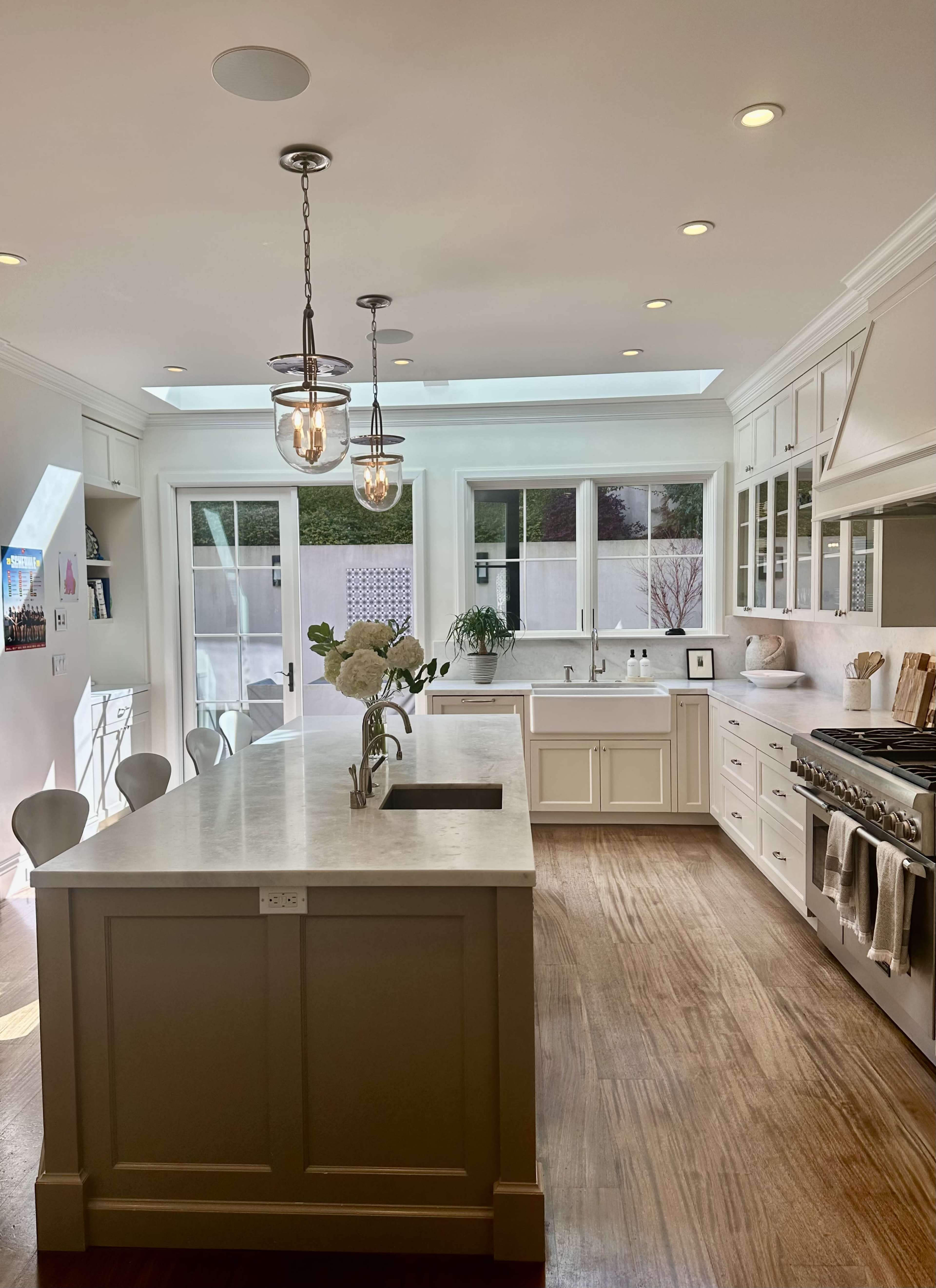 The image shows a modern kitchen with a central island, white cabinetry, and large windows allowing natural light to illuminate the space.