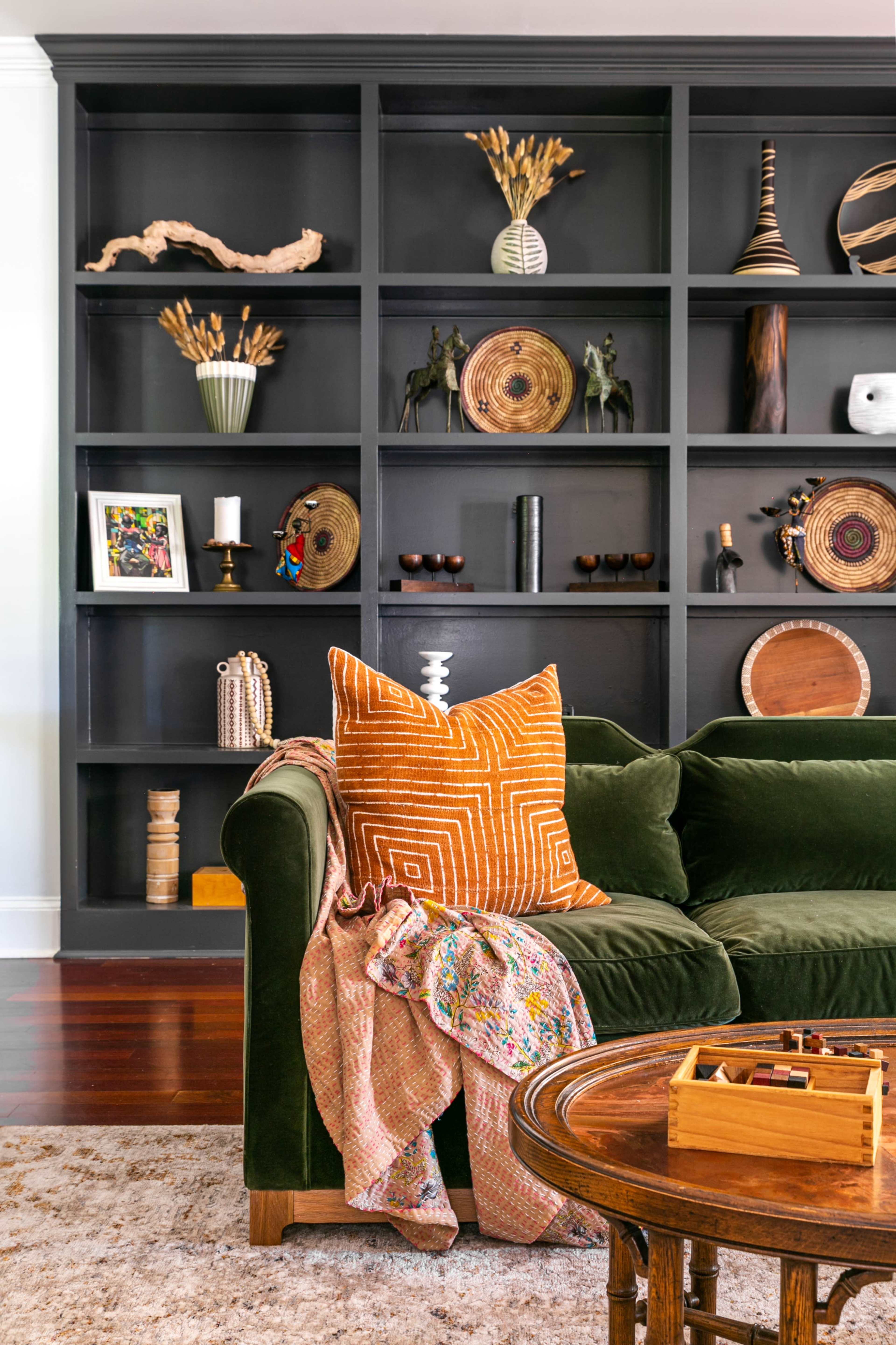 A green sofa with a patterned throw pillow and blanket is positioned in front of a dark shelving unit filled with various decorative items and artworks.