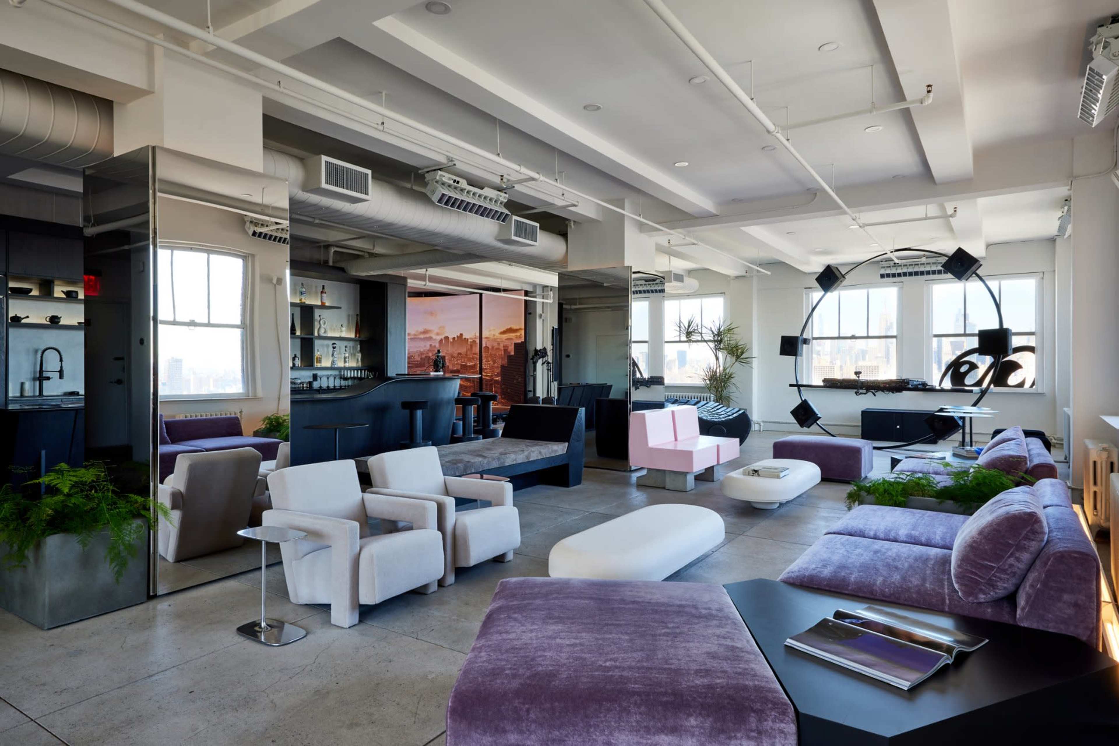 The image shows a modern, open-concept lounge area featuring various seating options, large windows with city views, and a stylish bar in the background.