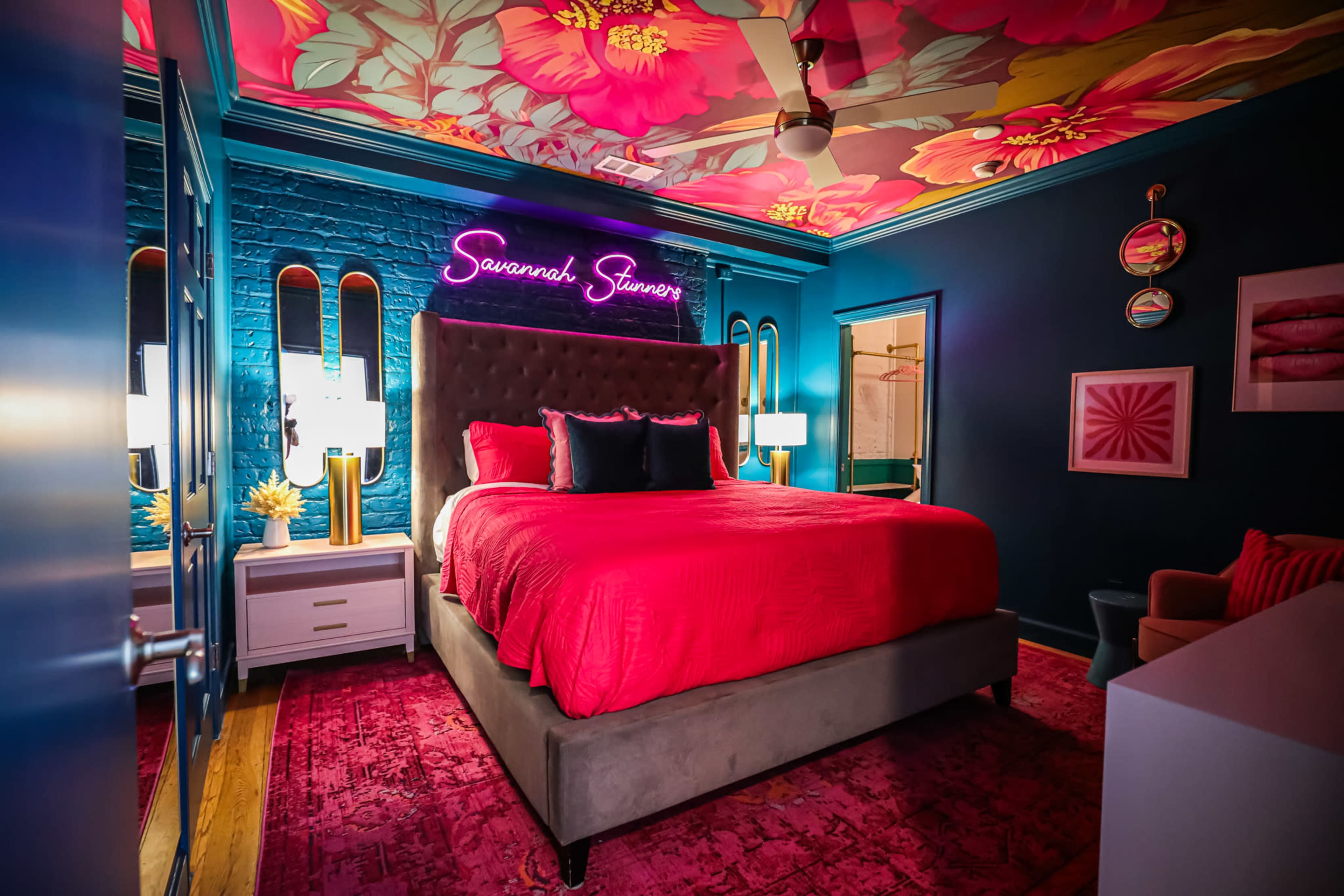 The bedroom features a large gray upholstered bed with bright red bedding, flanked by white bedside tables and illuminated by neon signs against a floral ceiling mural.