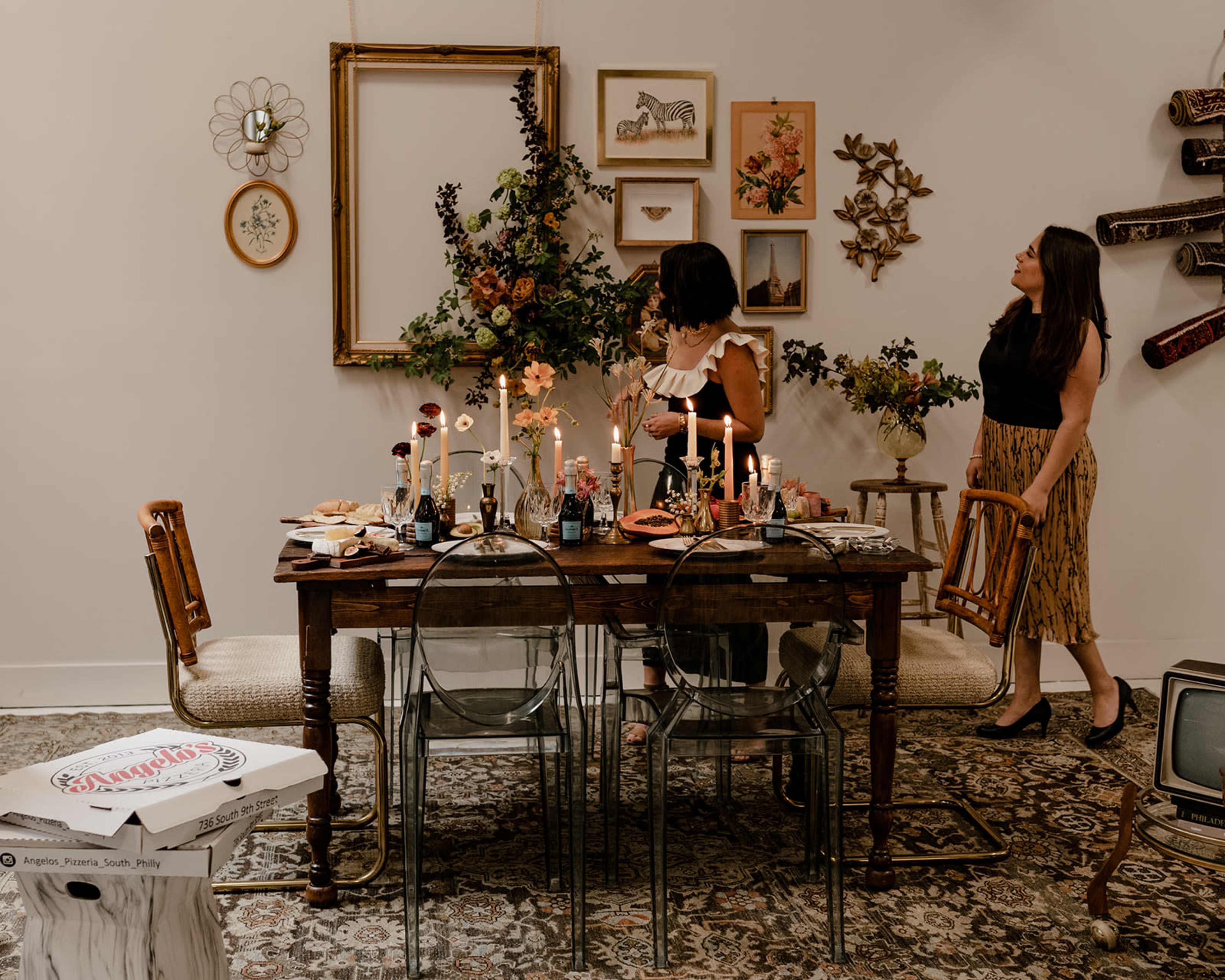 Two women stand in a dining area adorned with a table set for a meal, featuring candles, food, and floral decorations, while art pieces hang on the wall behind them.