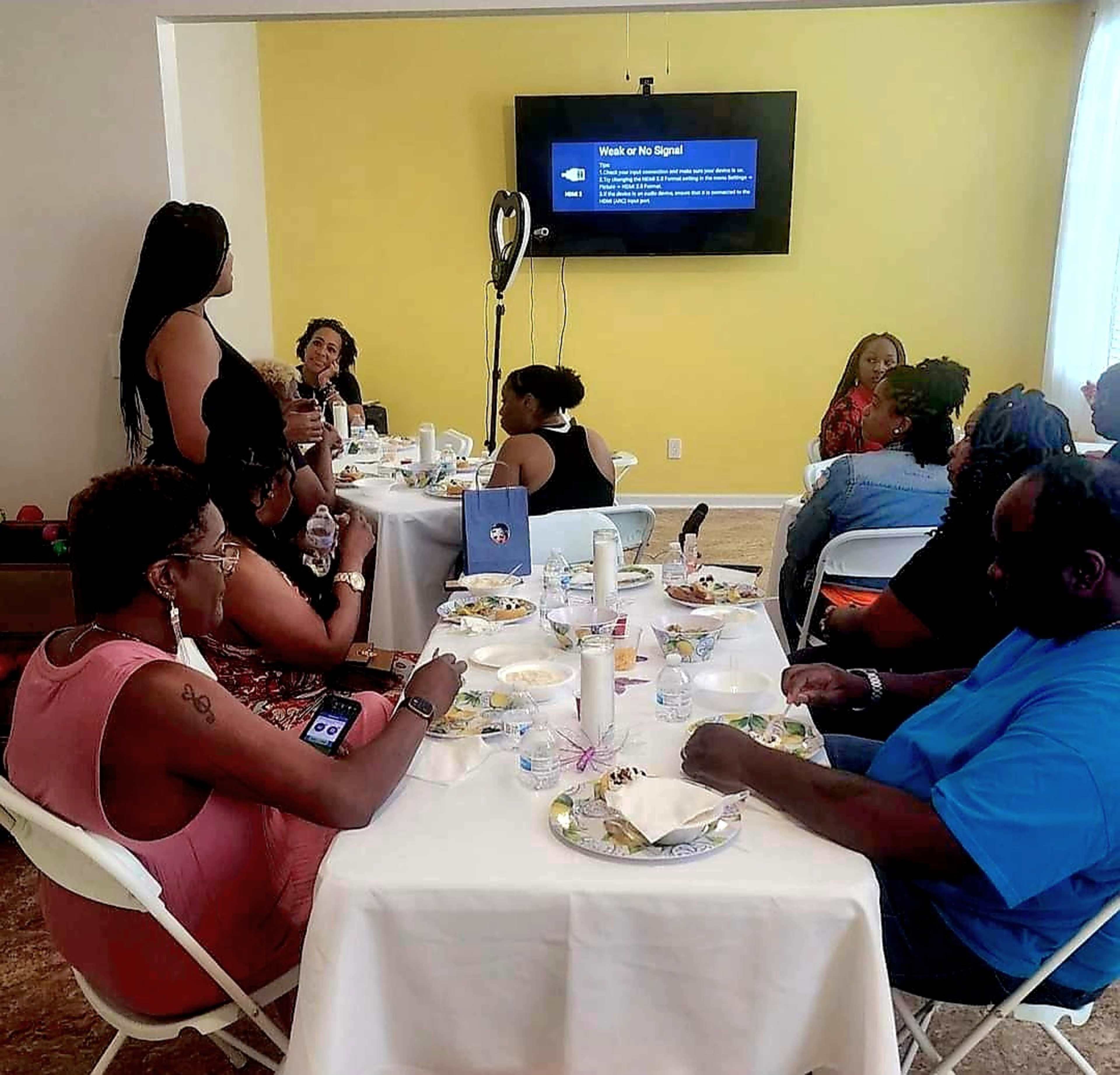 A group of people sits around tables sharing a meal while a woman stands and addresses the audience in a brightly lit room.