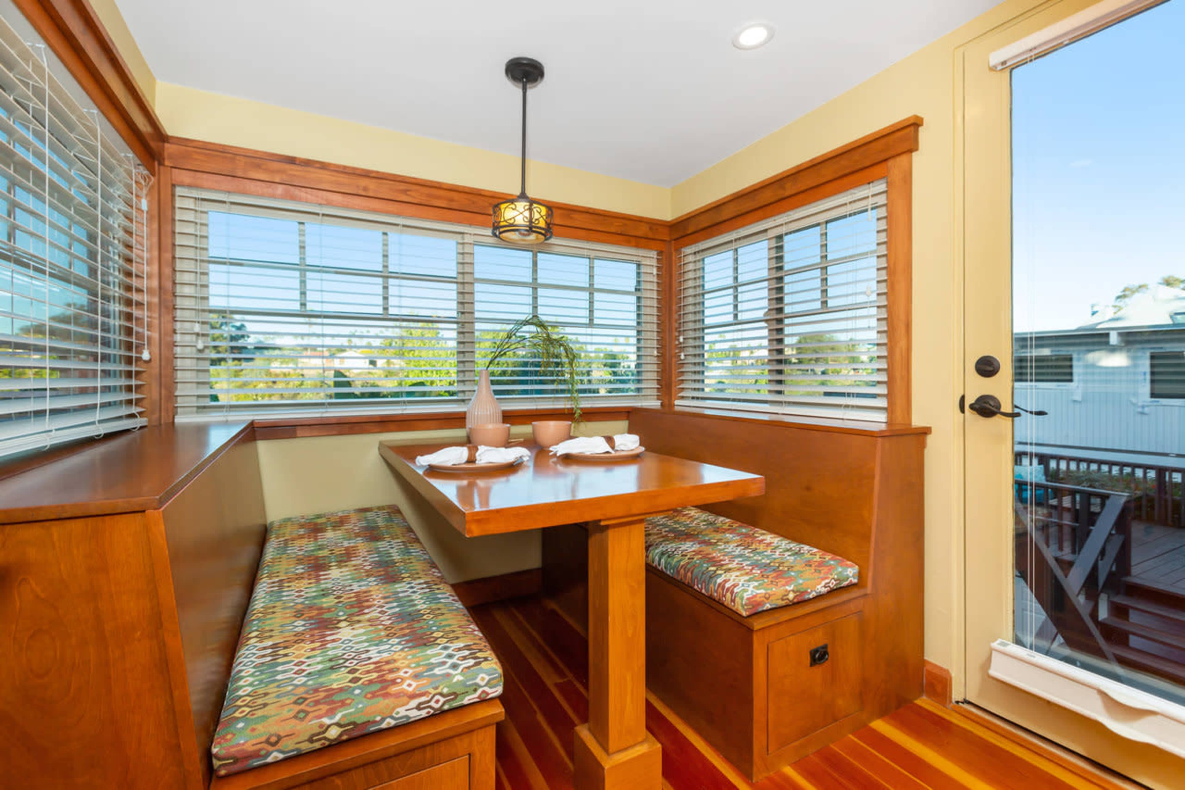 A cozy dining nook features a wooden table with patterned benches and large windows overlooking an outdoor deck.