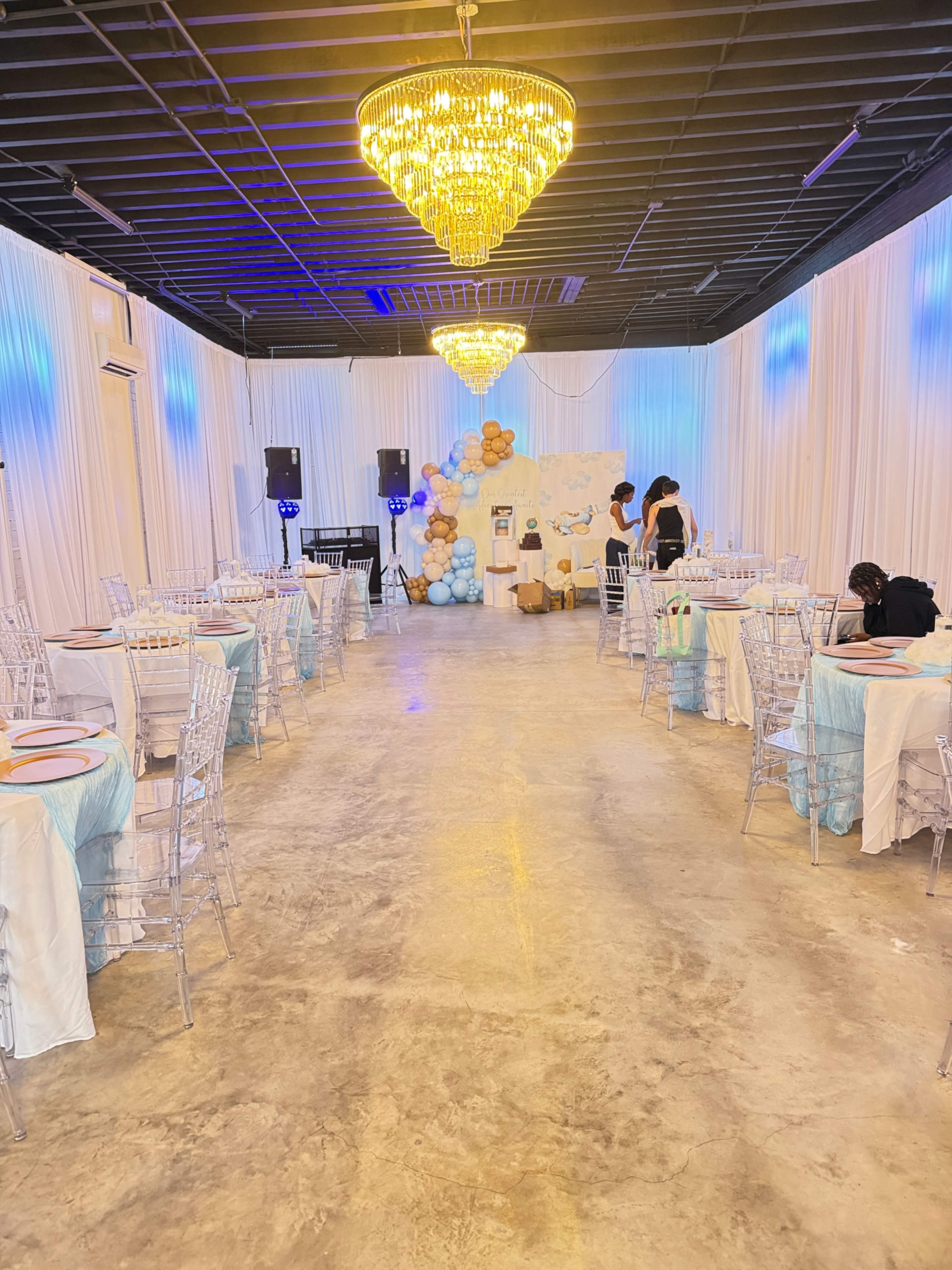 The image shows a spacious event venue decorated with white drapes, a backdrop of balloons, and several round tables set with light blue tablecloths and clear chairs.
