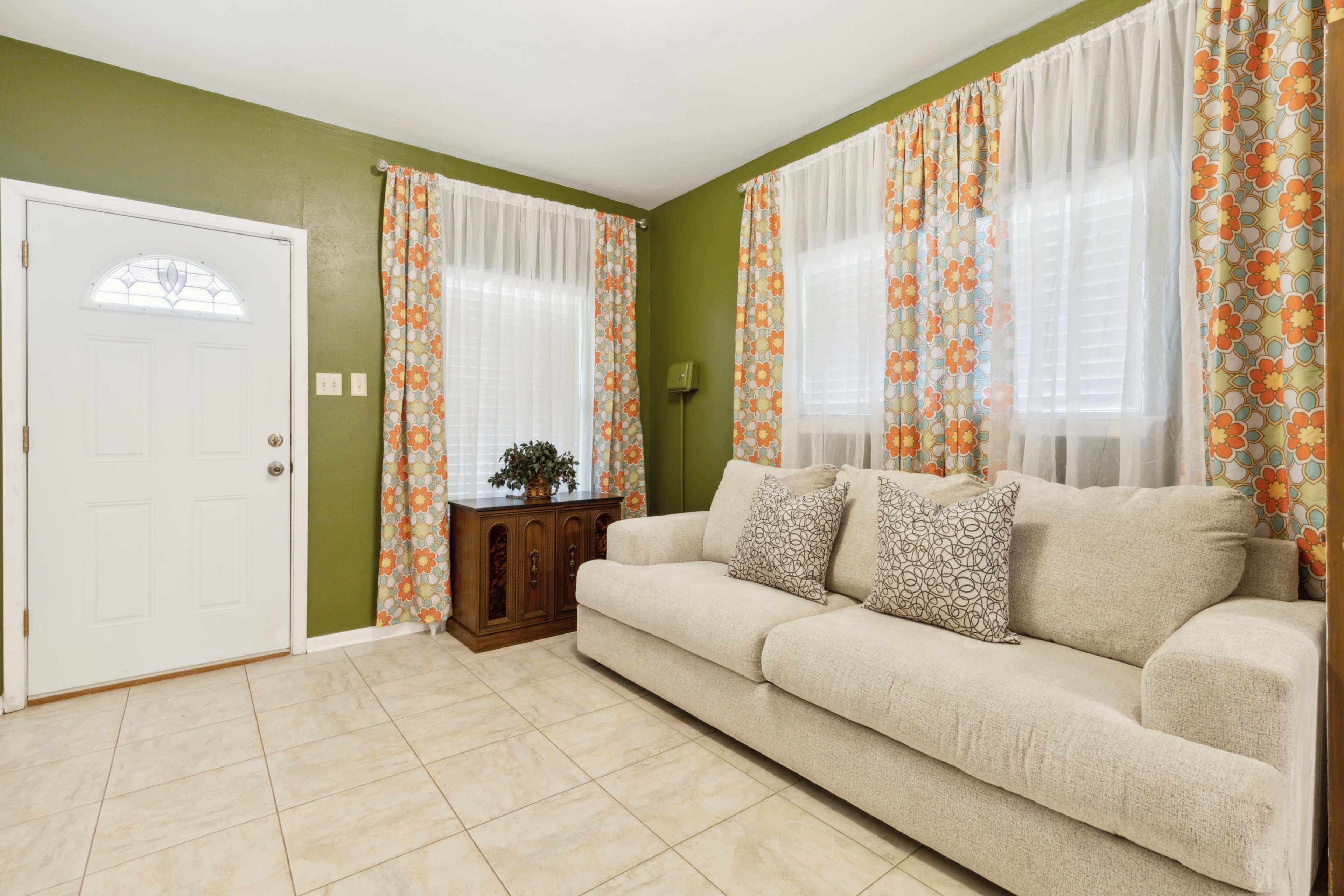 A light-colored sofa with decorative pillows is positioned in a room featuring green walls, large windows covered with floral curtains, and a wooden cabinet near the entrance.