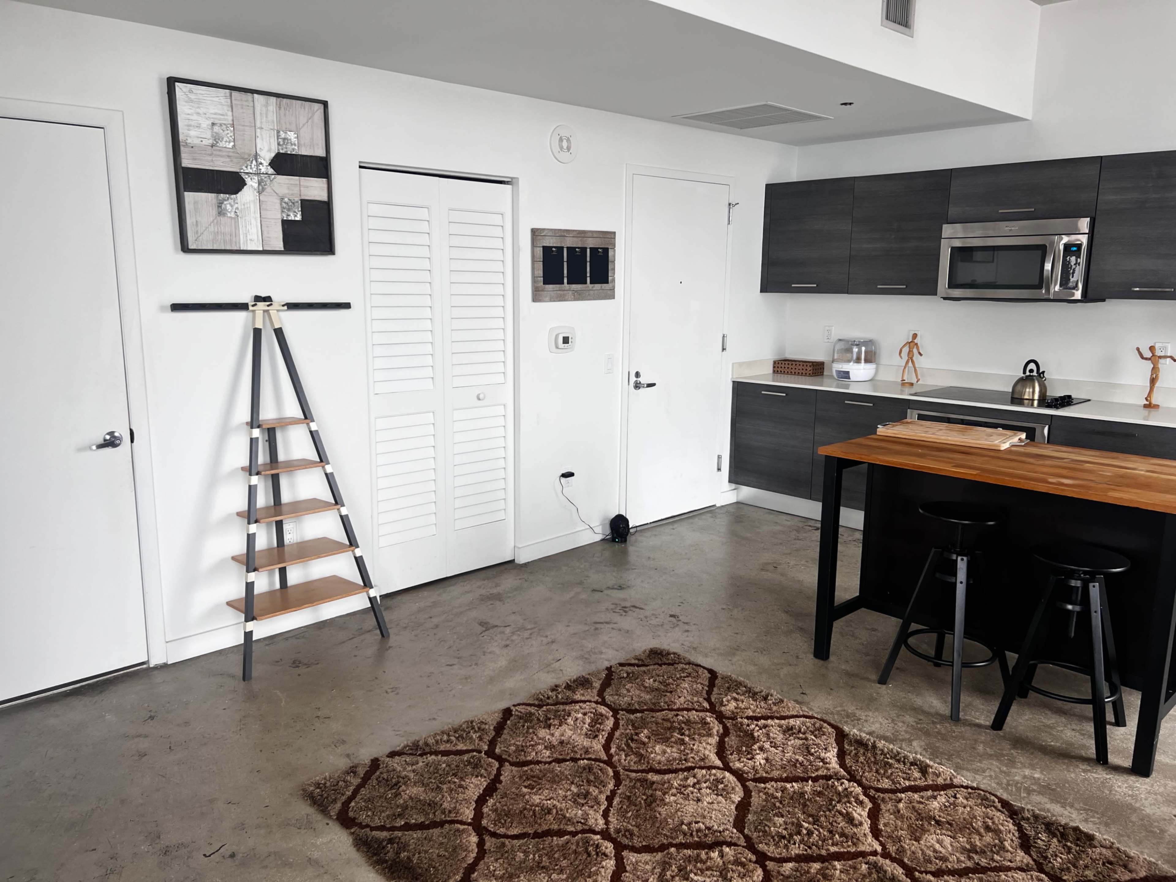 A modern kitchen and living space features a wooden island, black stools, and a decorative ladder beside a white door.