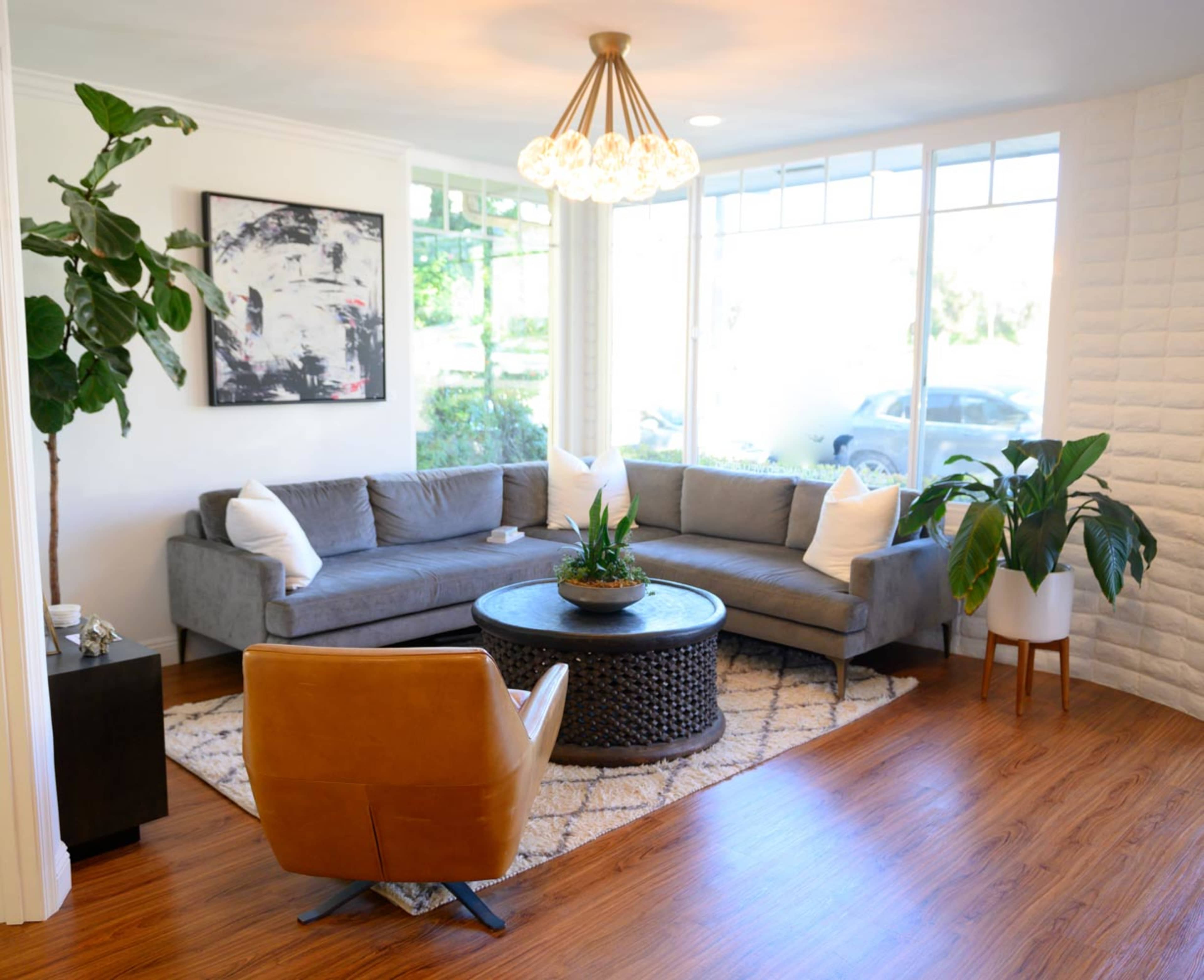 A modern living room features a gray sectional sofa, a round coffee table with greenery, and large windows allowing natural light.