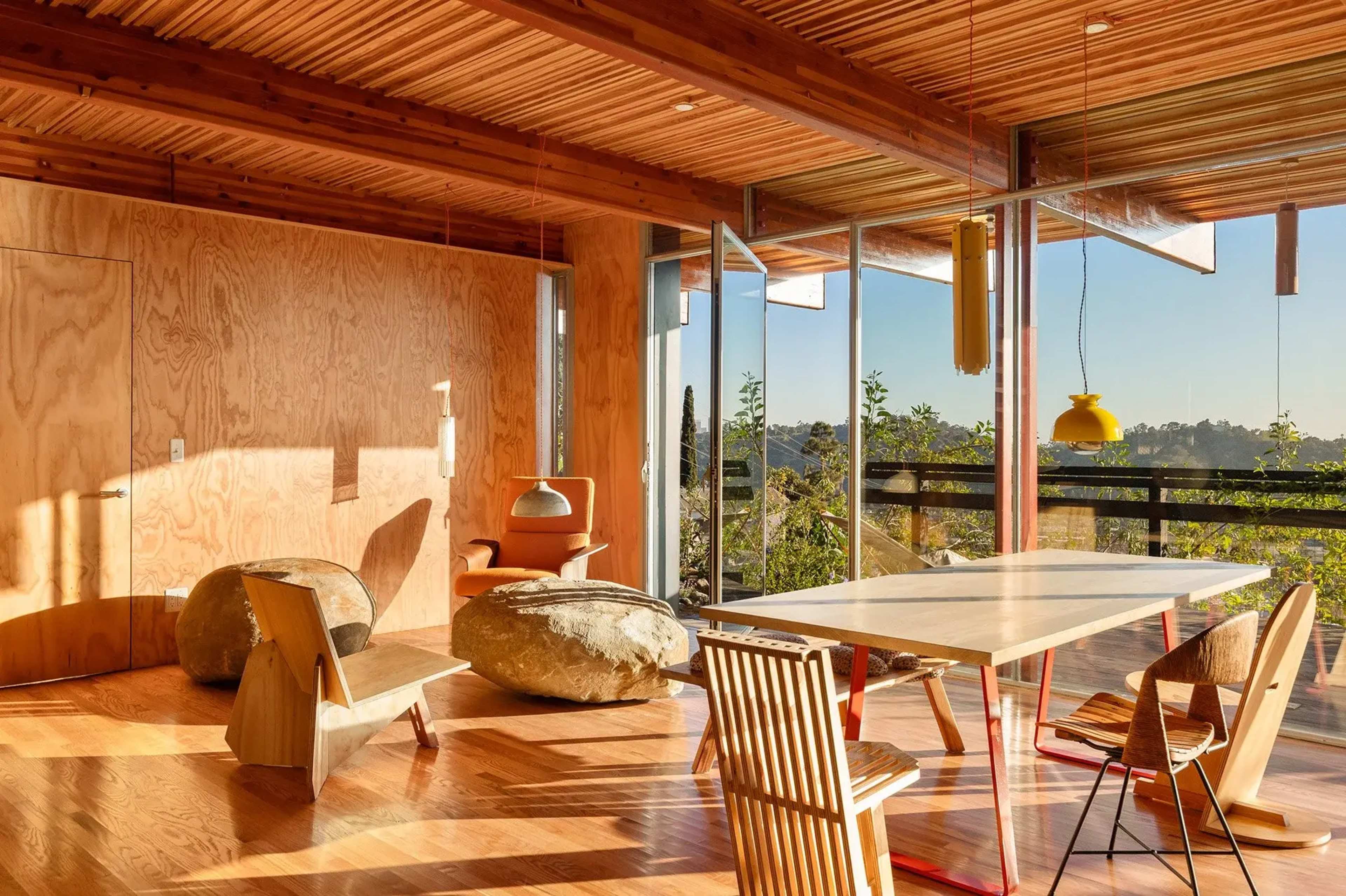 The image shows a modern interior space with wooden walls, large windows, and a dining area featuring a light-colored table and several chairs.