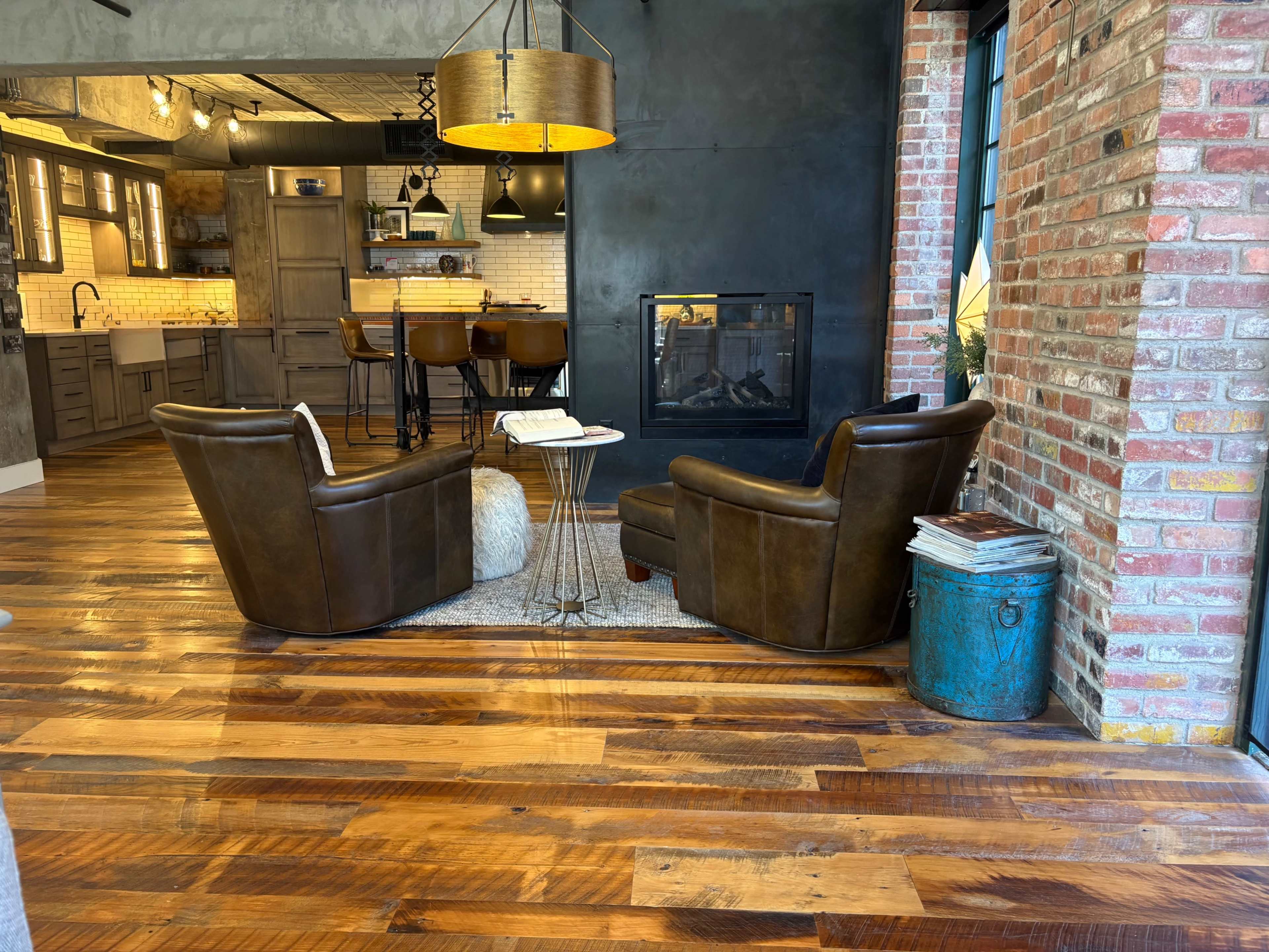 The image features a cozy seating area with two leather armchairs facing a fireplace, set against a backdrop of a modern kitchen with exposed brick walls and wooden flooring.
