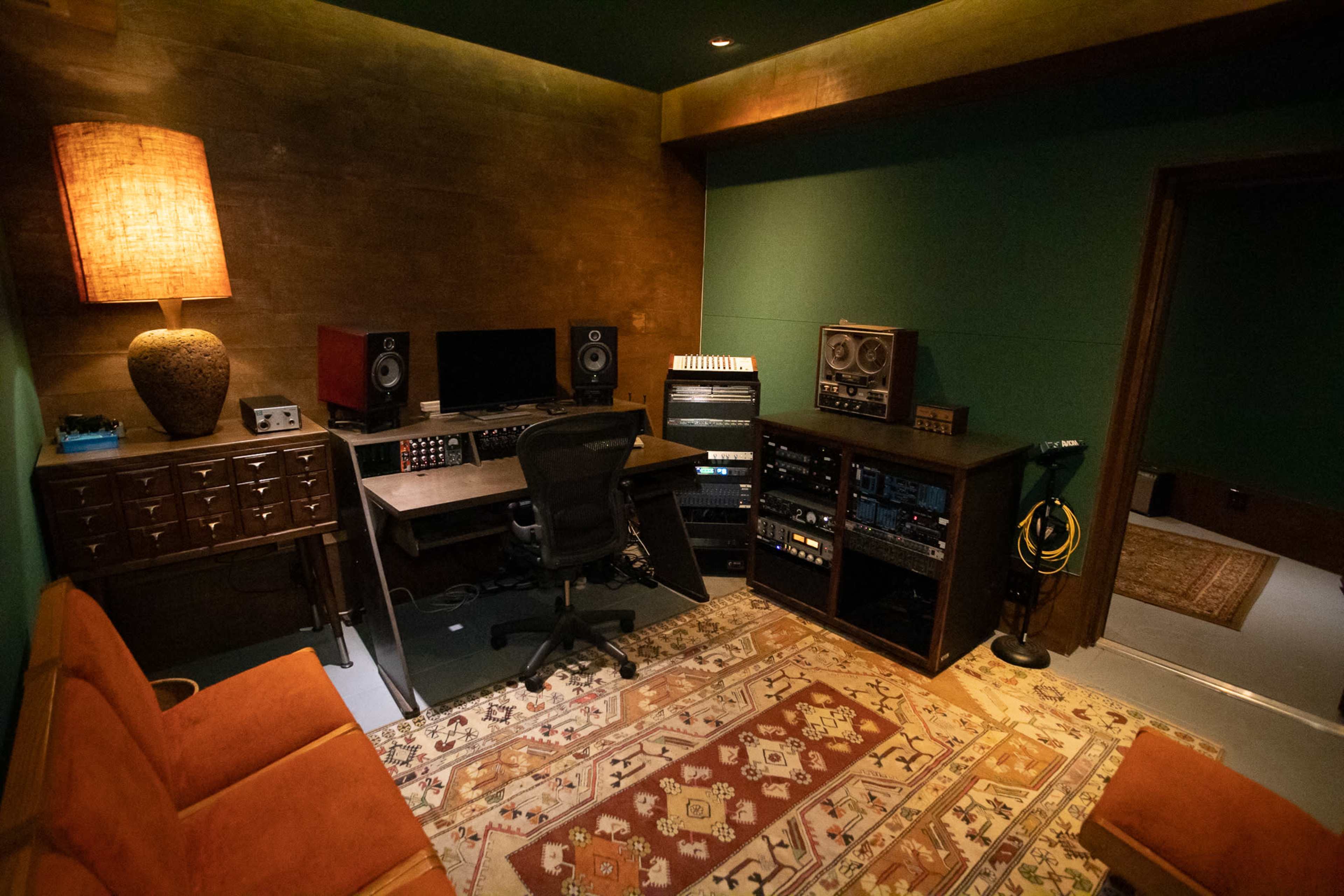 The image depicts a small studio room featuring a desk with computer monitors, audio equipment, and speakers, along with a seating area and a patterned rug.