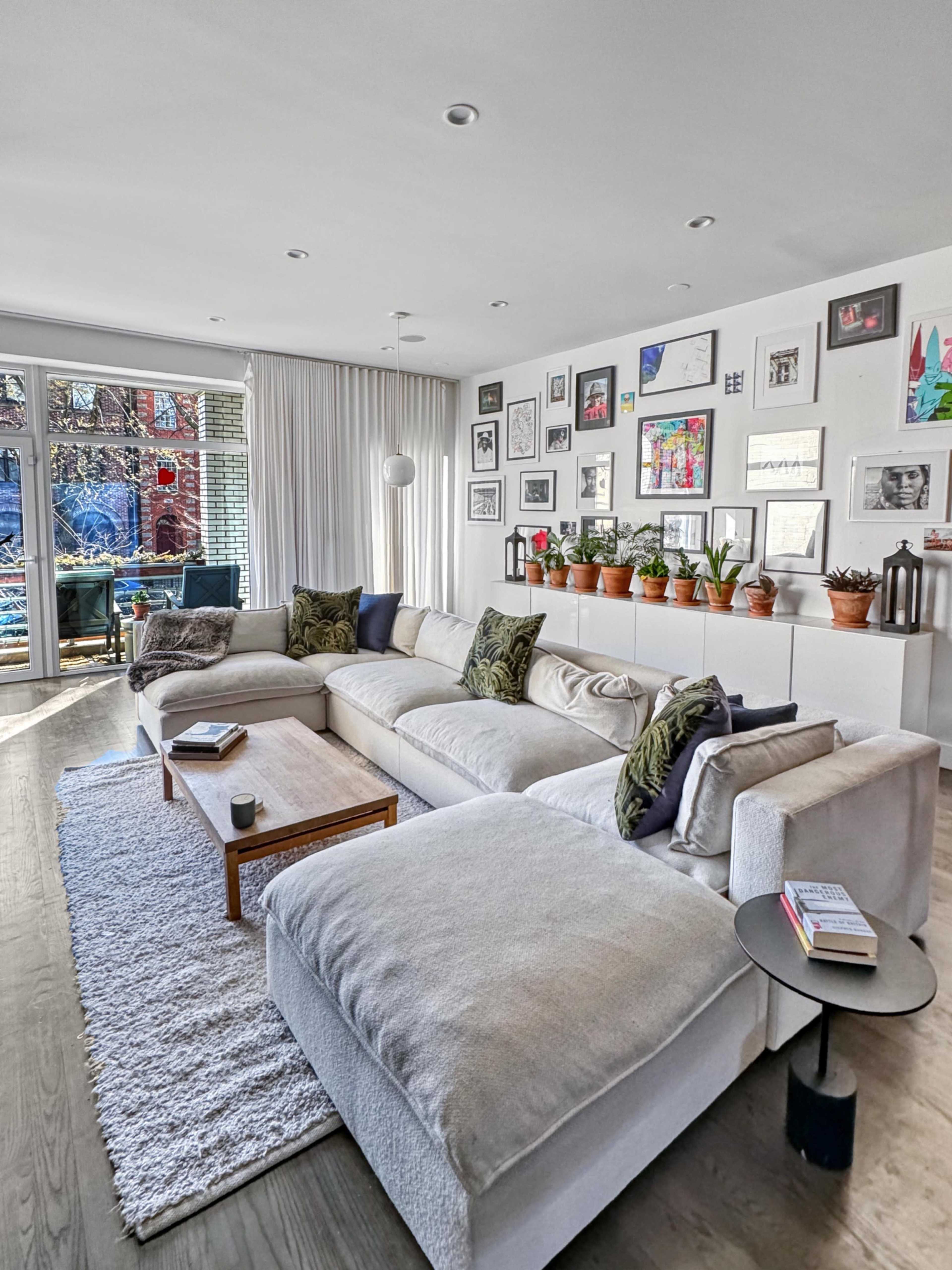 A spacious living room features a large sectional couch, a coffee table, and walls adorned with framed pictures and potted plants.
