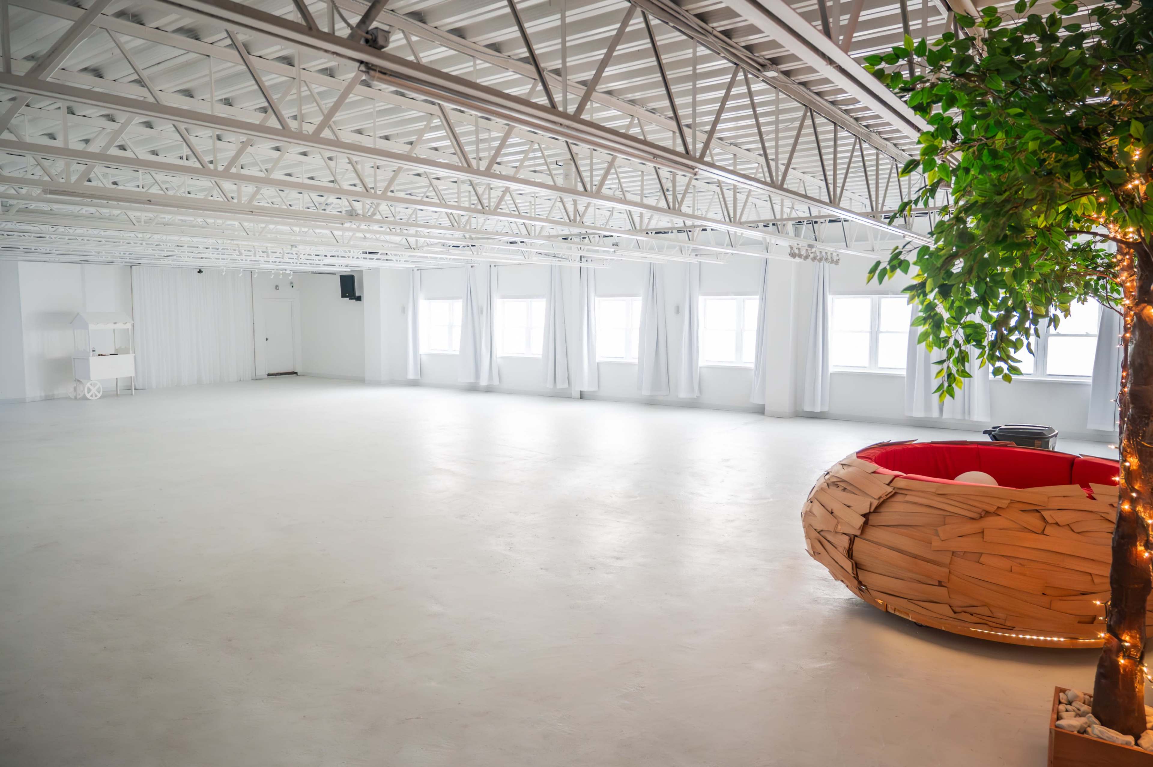 The image shows a spacious, empty room with white walls, large windows, and a circular wooden seating area filled with red cushions.