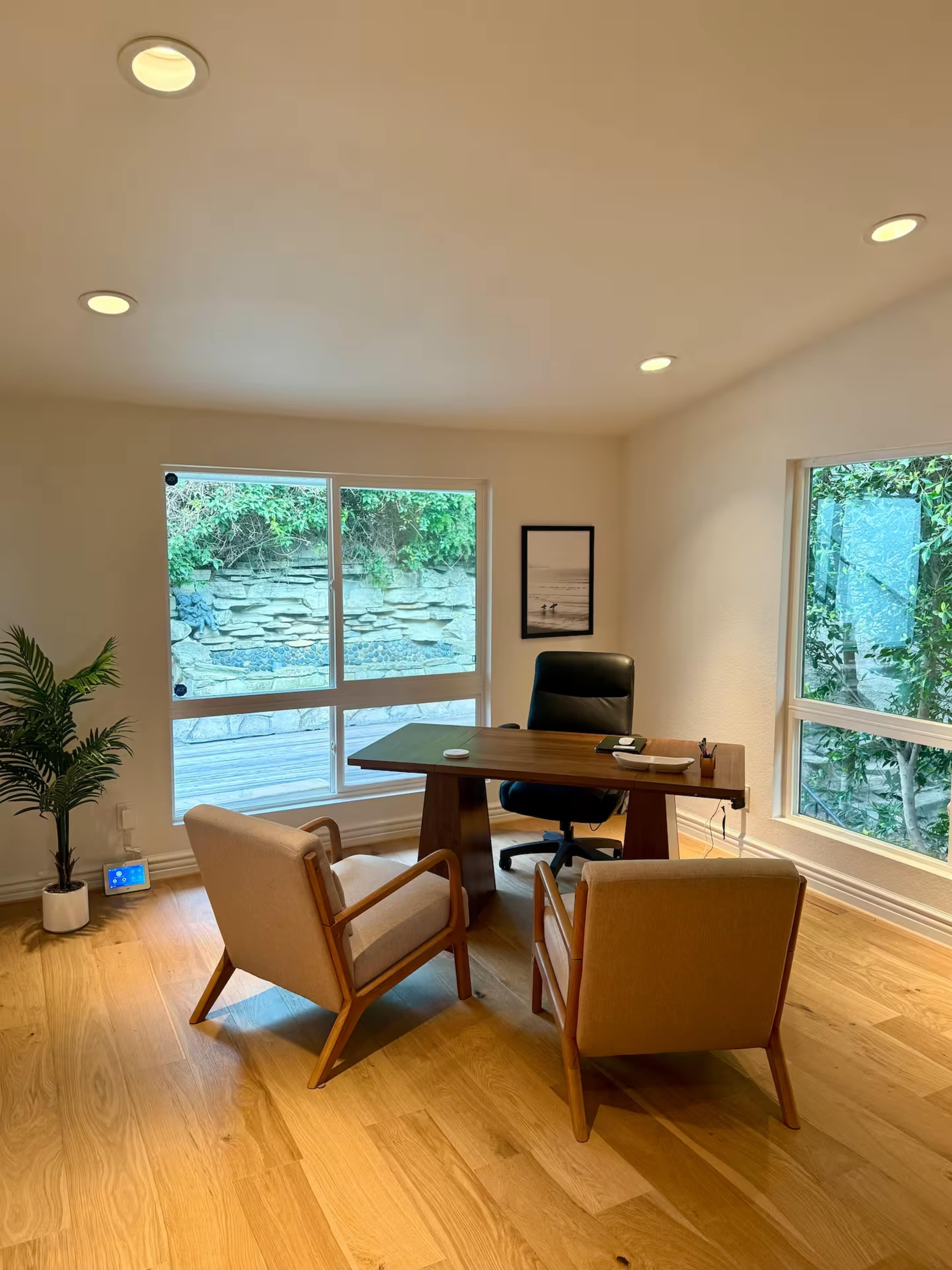 The image shows a modern office space featuring a wooden desk, two armchairs, and a large window facing a green outdoor area.