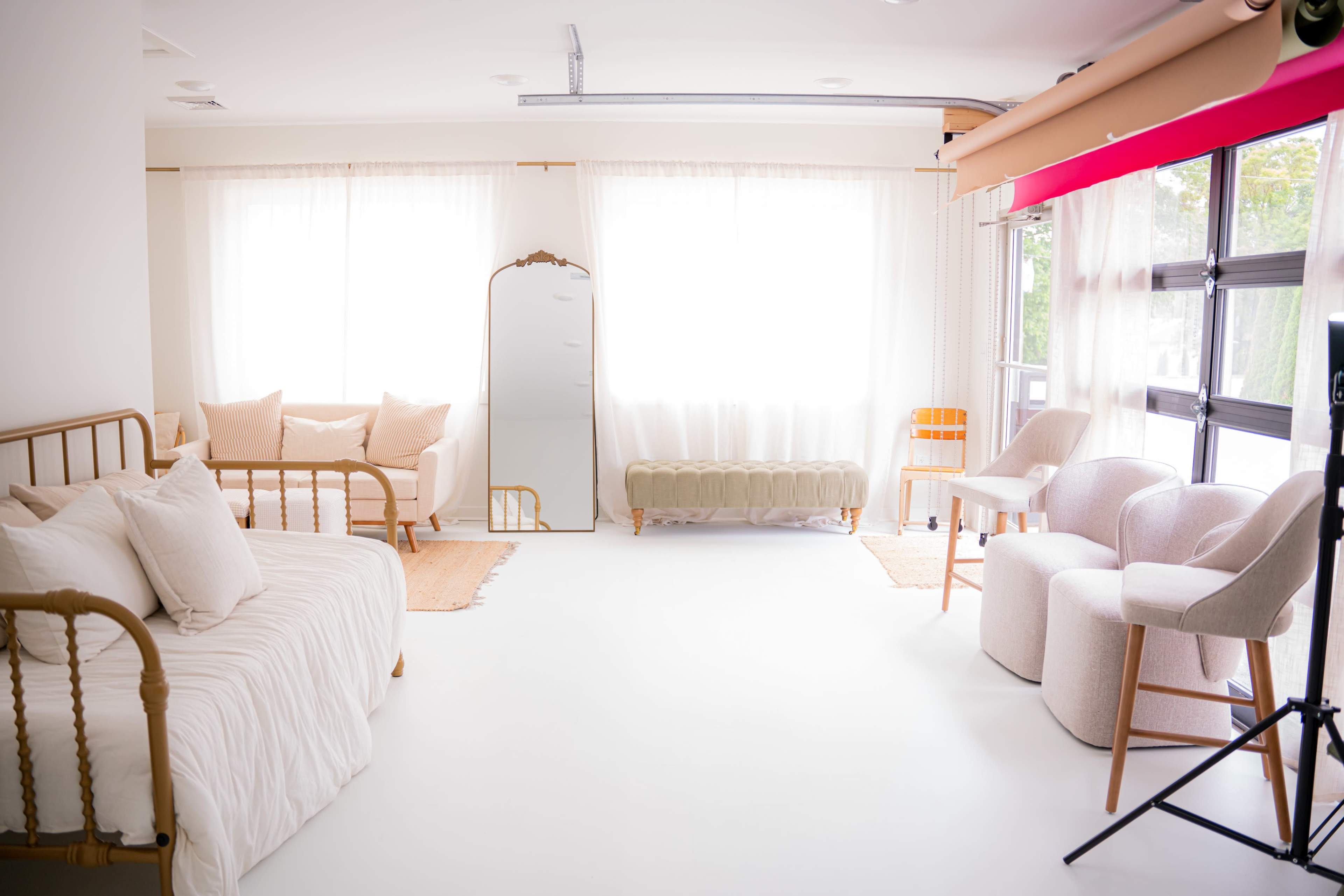 The image shows a bright, minimalistic interior space with a daybed, two armchairs, a mirror, and a green upholstered bench, all set against large windows with sheer curtains.