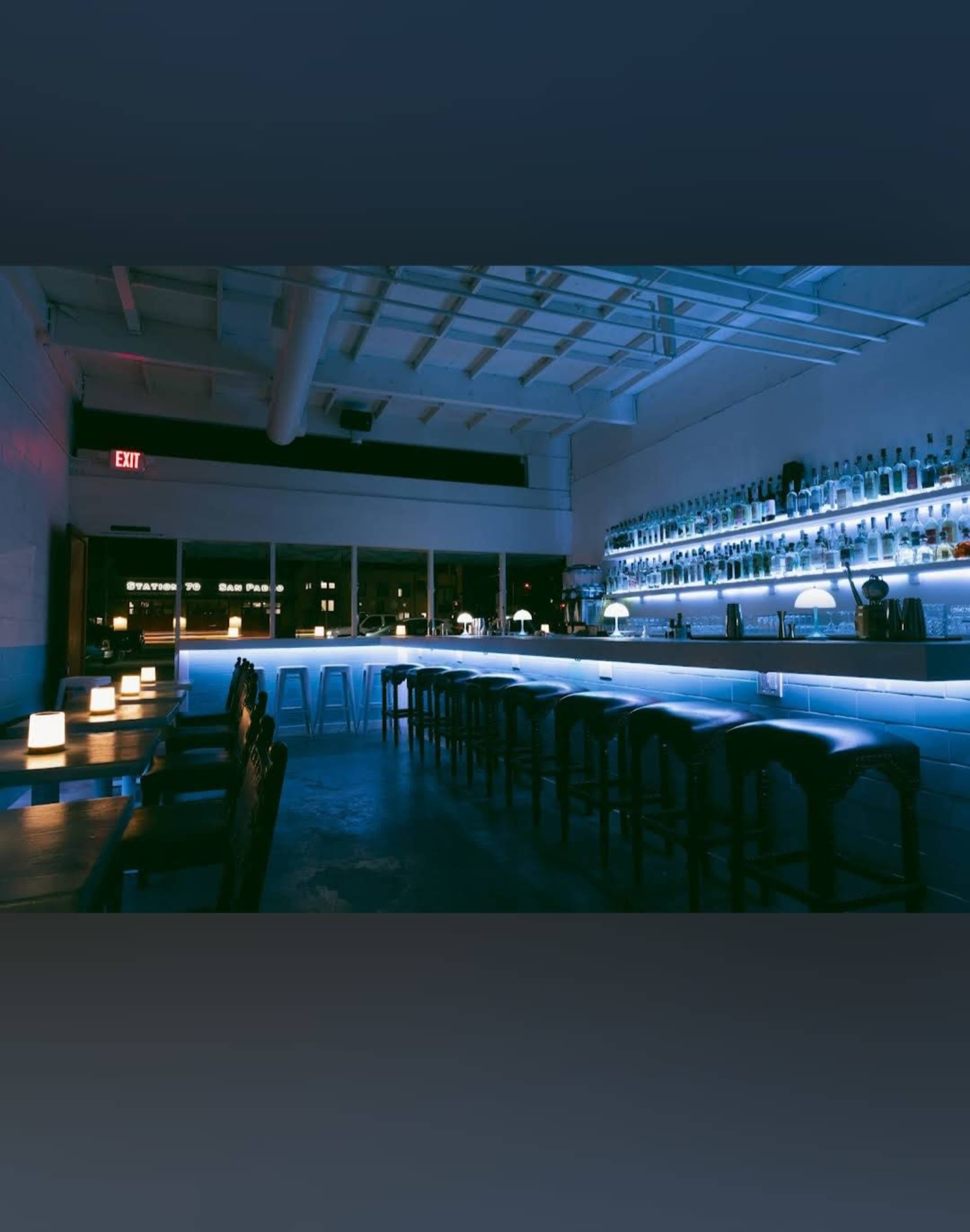 A well-lit bar features a long countertop with stools, illuminated by blue lights and surrounded by shelves of liquor bottles.