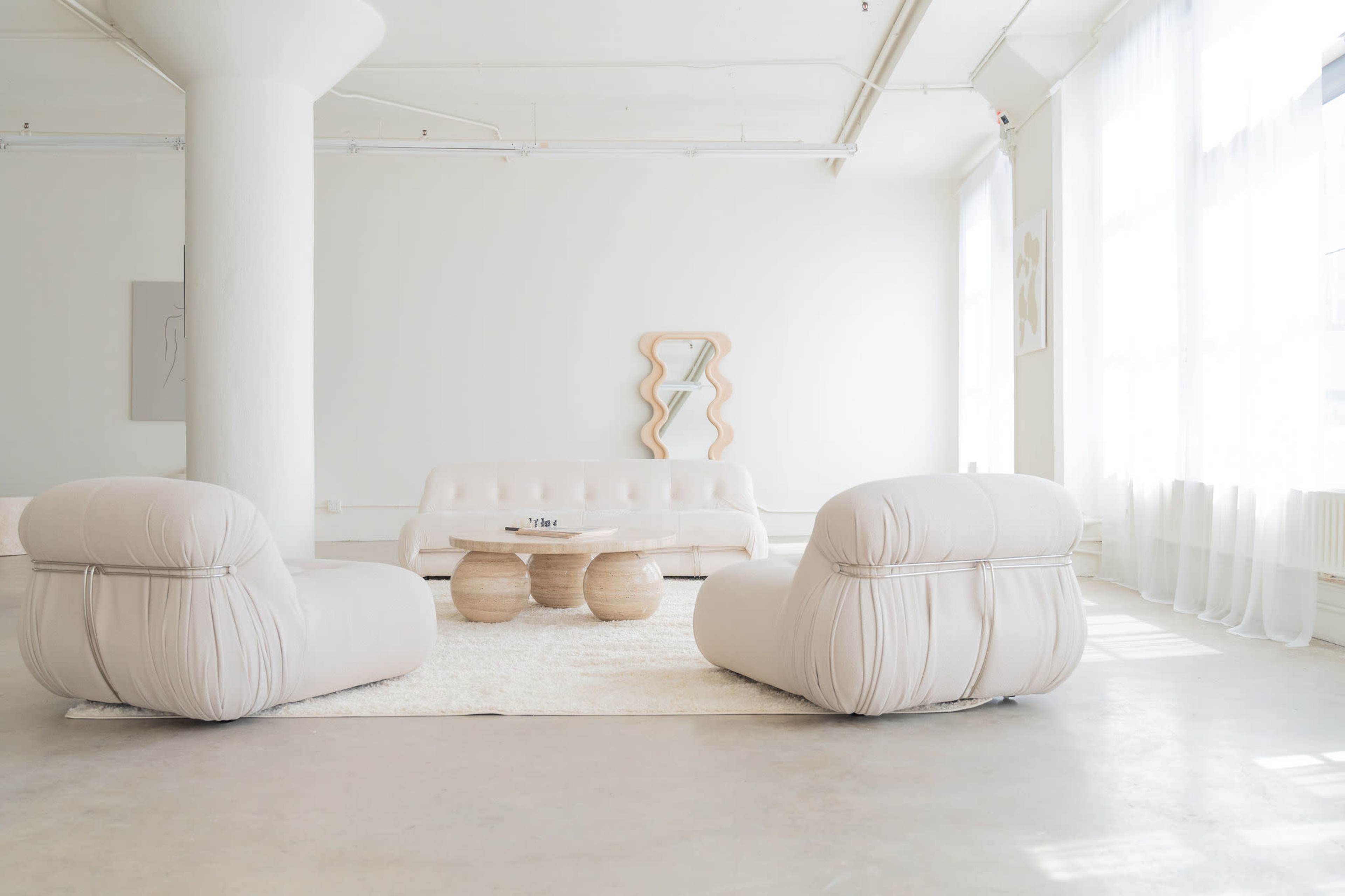 A minimalist interior featuring a light-colored couch, two round armchairs, a wooden coffee table, and large windows allowing natural light to fill the space.
