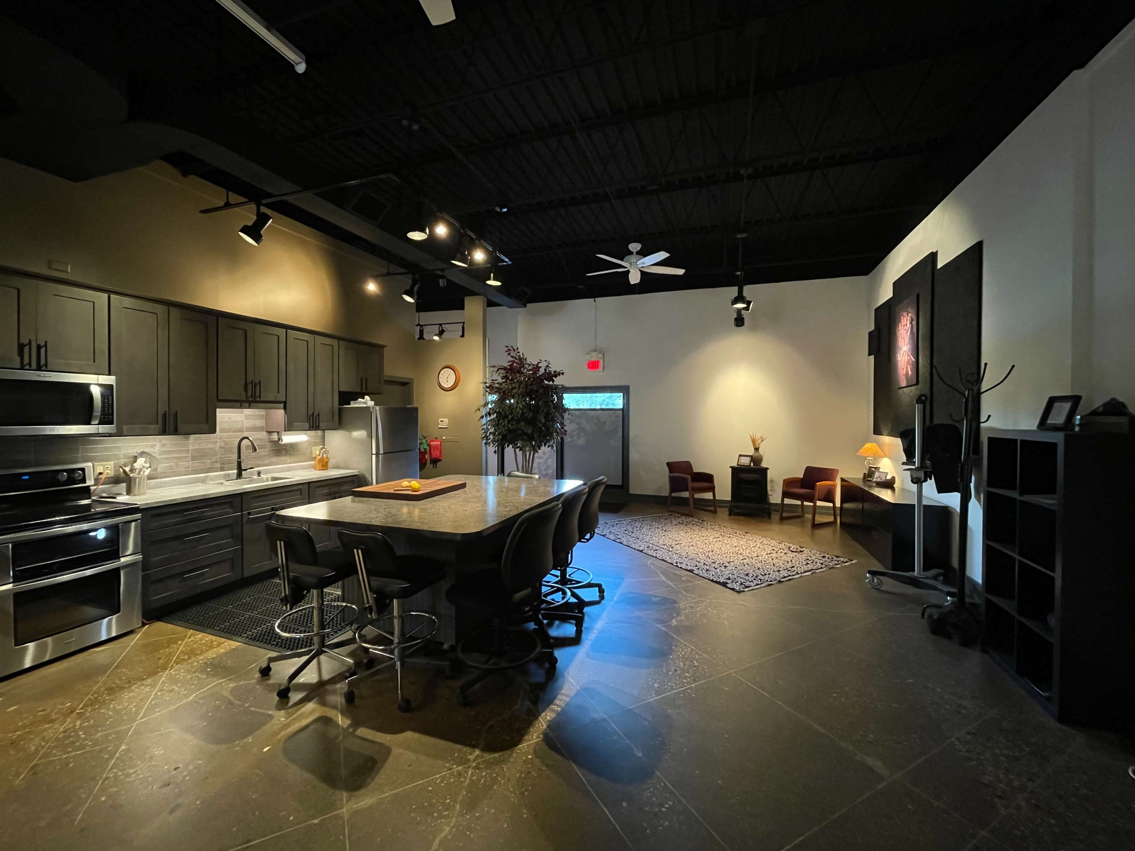 A modern kitchen with dark cabinetry, stainless steel appliances, and a large island, adjacent to a spacious living area with two chairs and a rug.