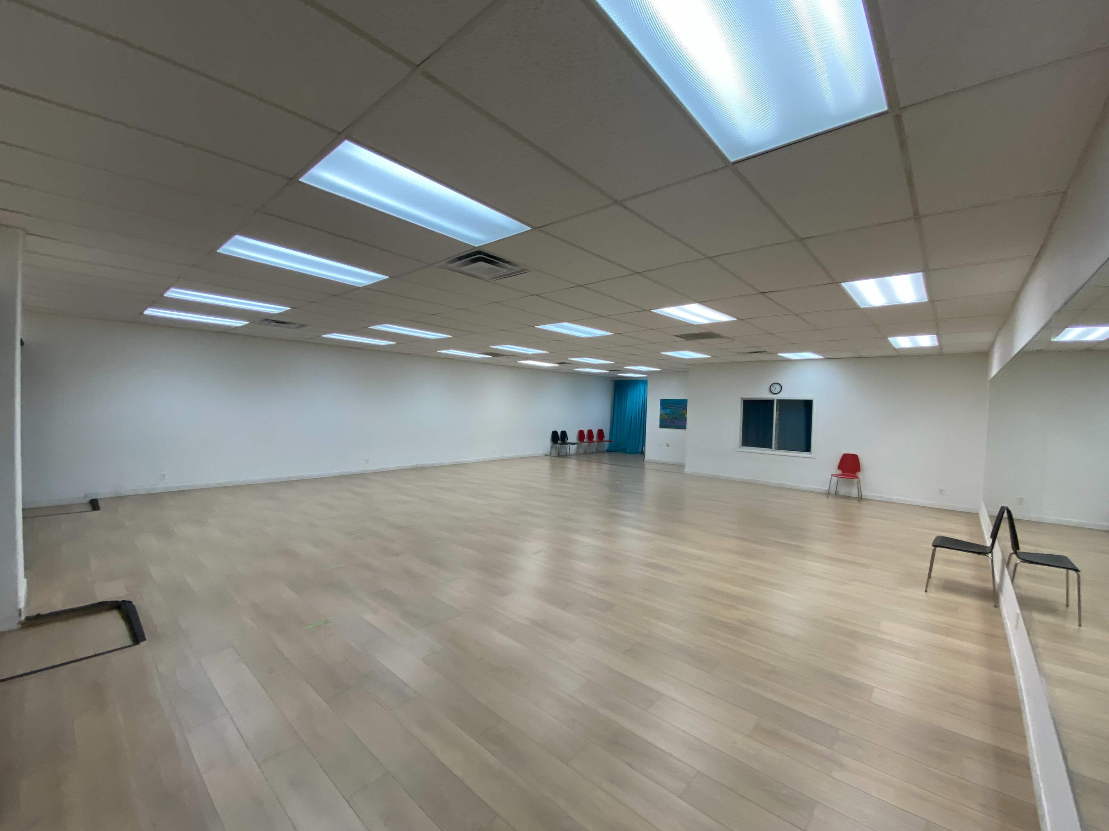 A spacious, empty room with wooden flooring, fluorescent lighting, and a large mirror along one wall.
