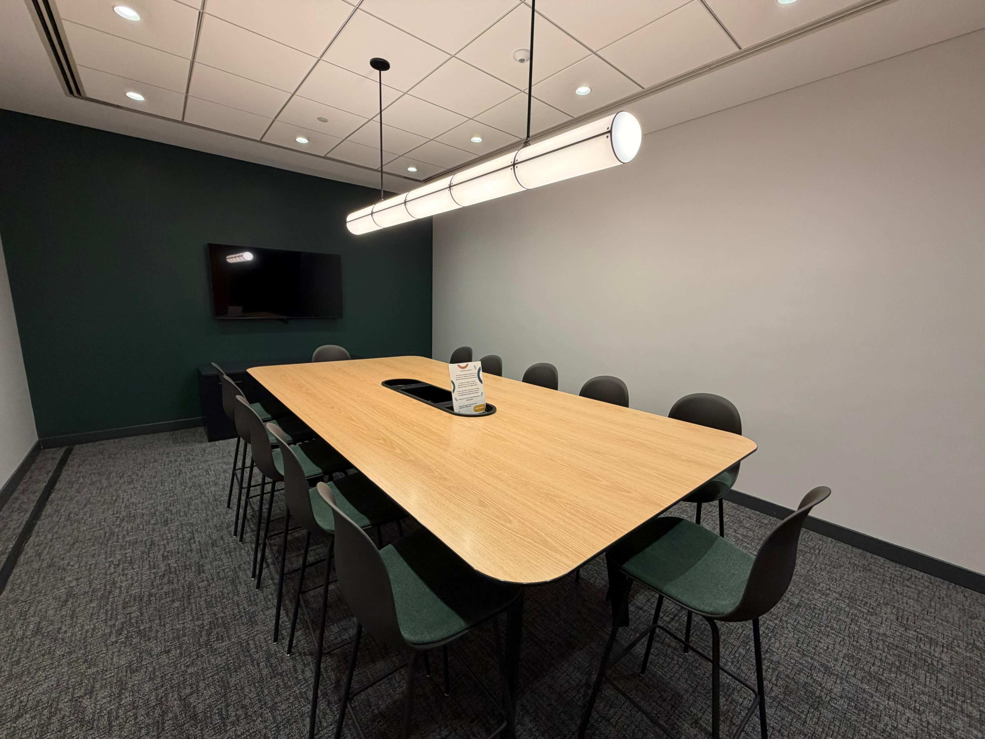 A modern conference room features a long wooden table surrounded by eight chairs, with a wall-mounted screen and overhead lighting.