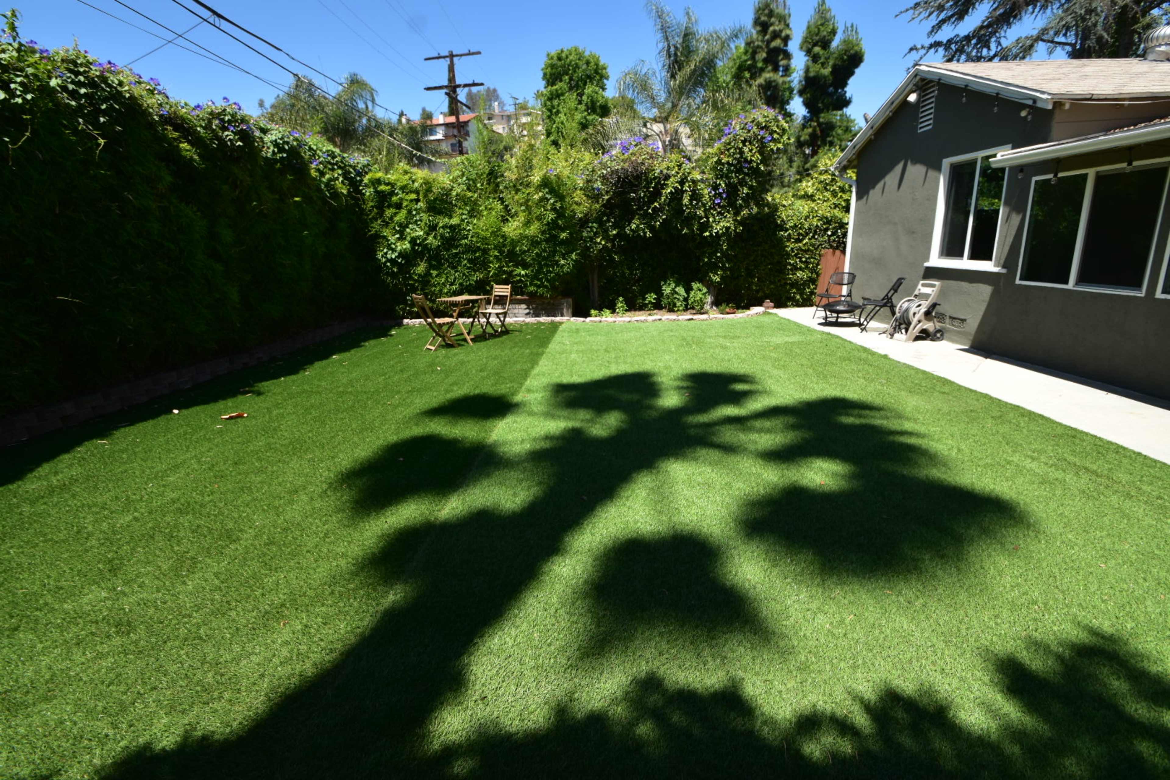 A green lawn with artificial turf features the shadow of a palm tree, surrounded by bushes and a grey house.