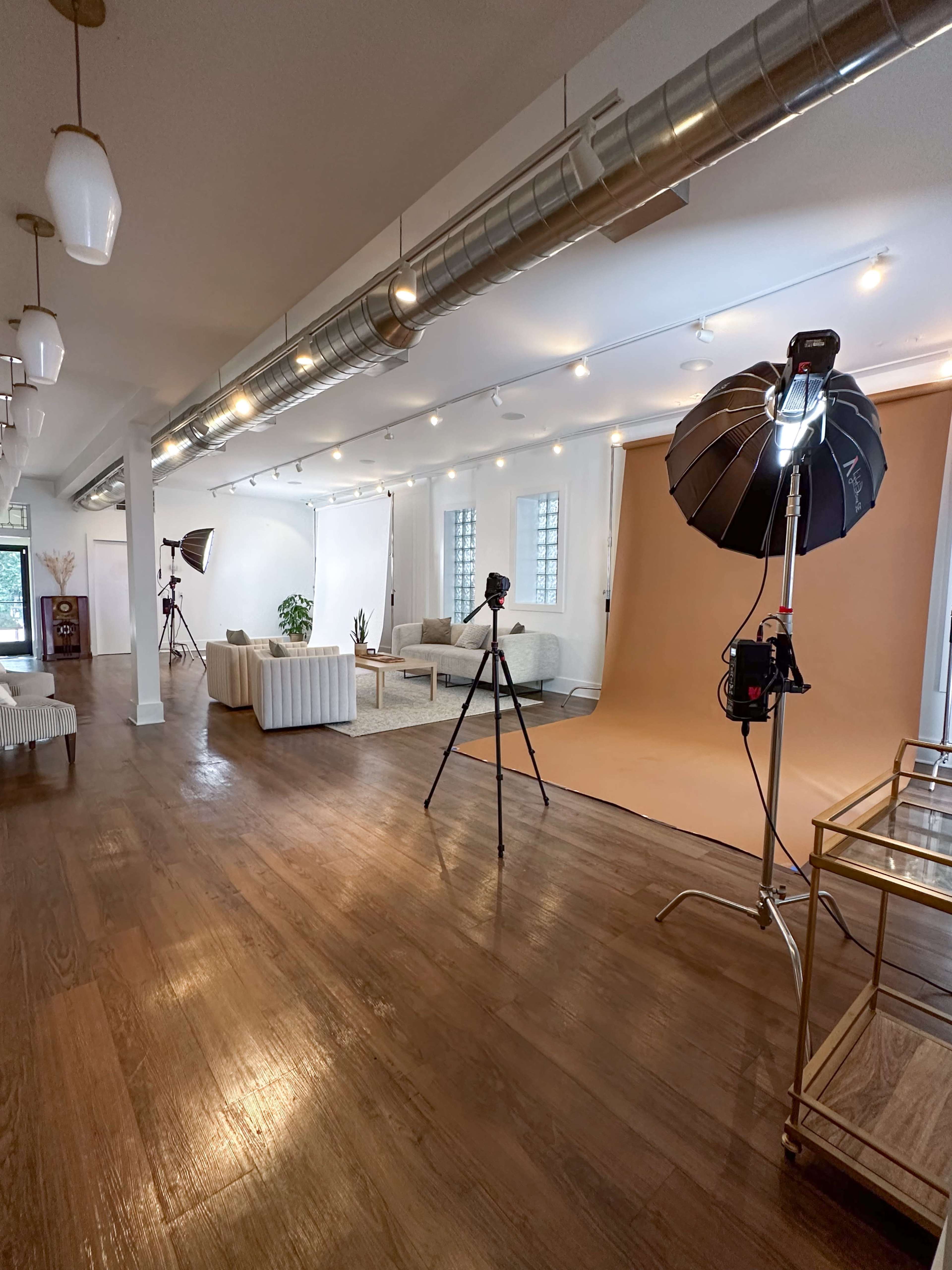 A photography studio features a light brown backdrop, soft lighting equipment, and a well-furnished seating area.